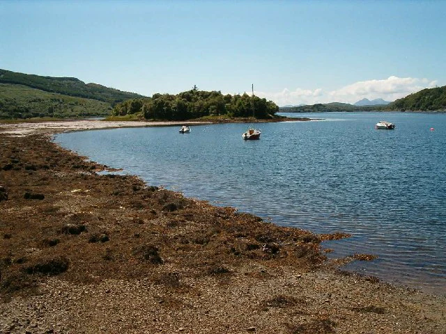 A wide angle view of the shoreline of Loch Sween with the island of Eilean Mhartan in the distance; three small boats are moored in the calm sea loch.