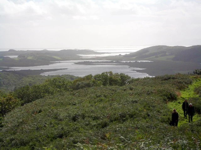A wide landscape view of Loch Sween in Scotland, showing the sea loch, its inlets, and surrounding hills, with a group of three people walking on a grassy trail in the foreground.