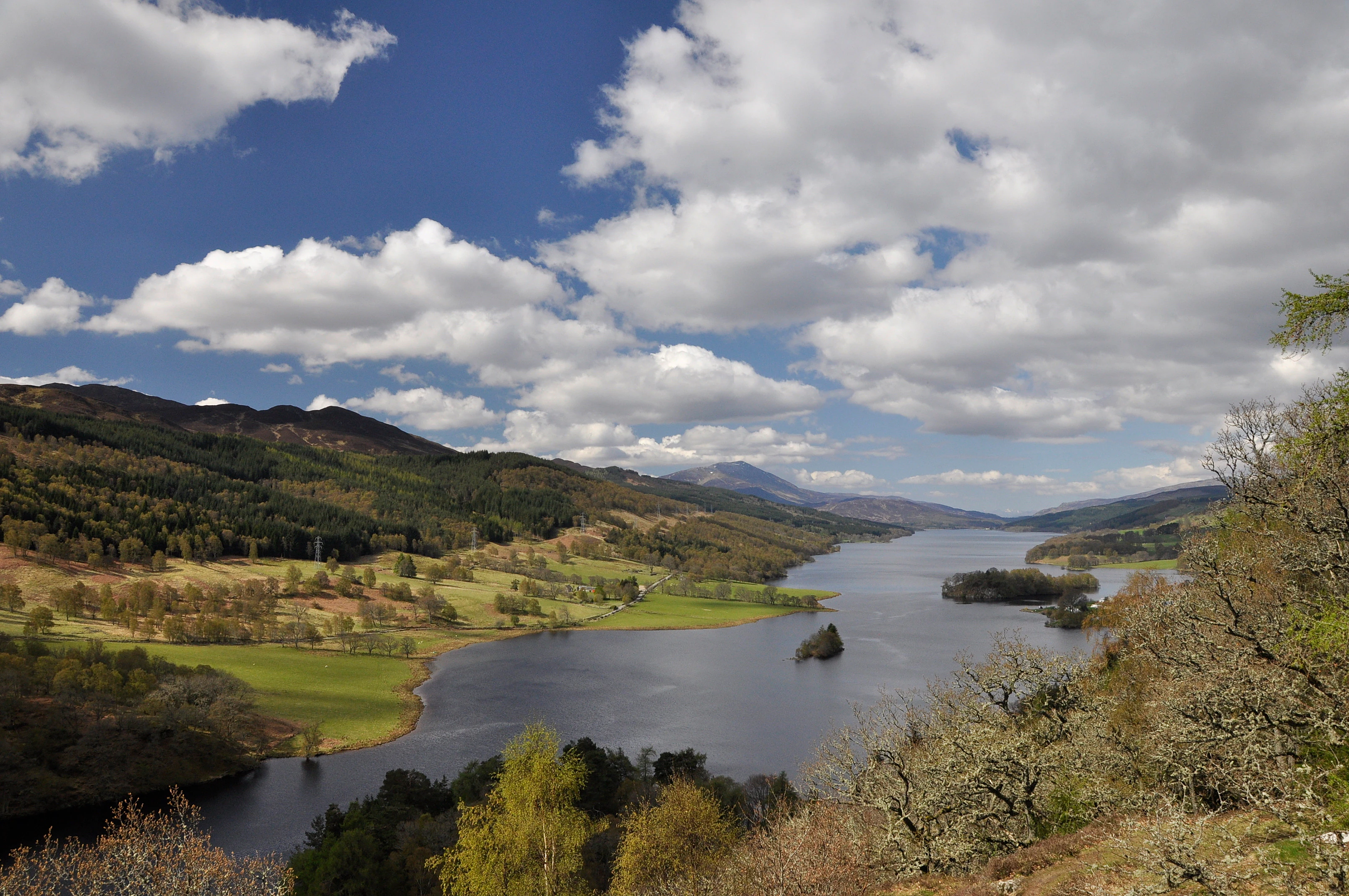 Una fotografía de paisaje gran angular que mira hacia el lago Tummel en Escocia desde un punto elevado, mostrando el lago largo y estrecho rodeado de colinas verdes onduladas, bosques y un cielo nublado azul.