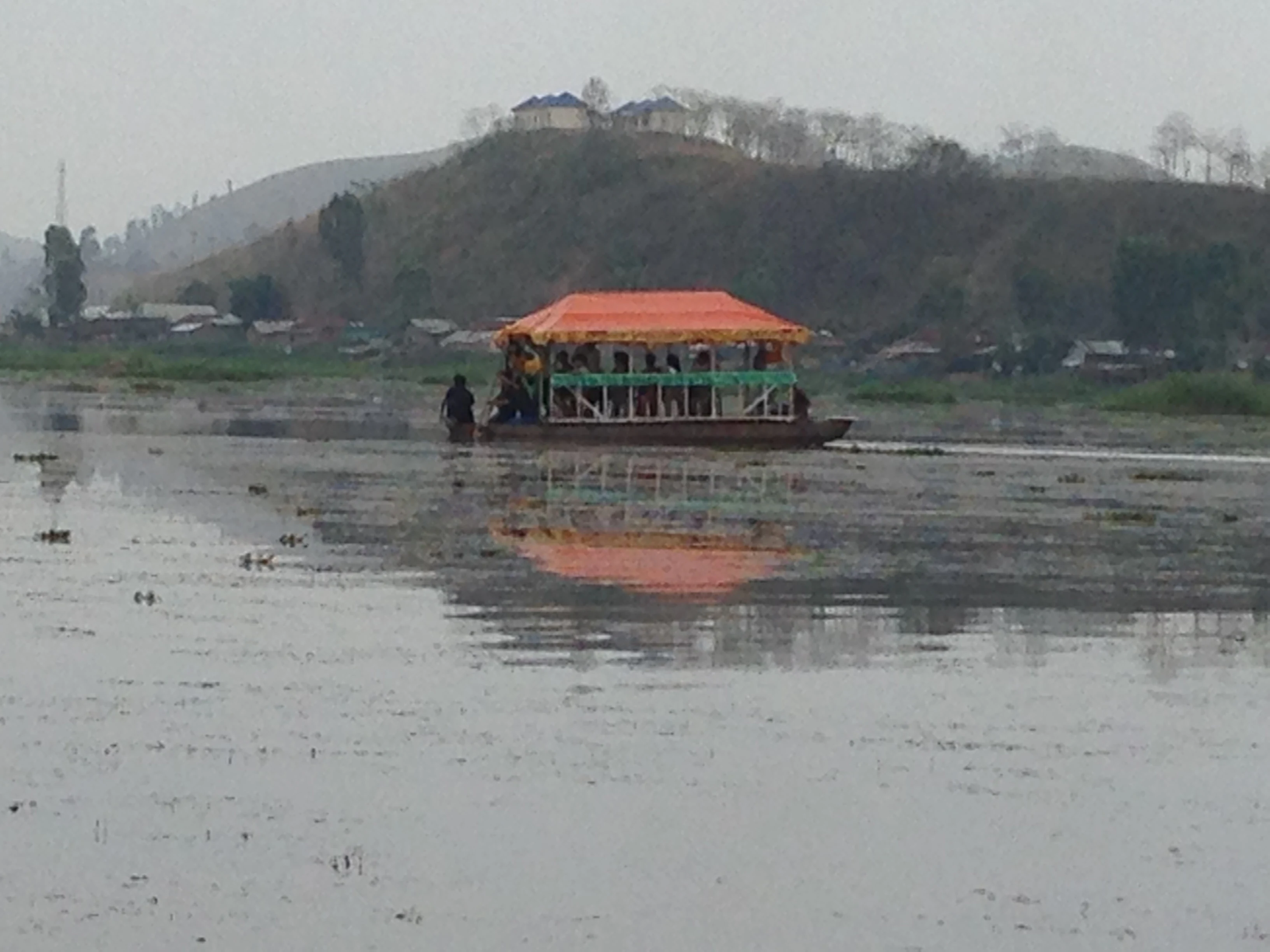 A passenger boat with an orange canopy traveling across a calm, flat, grey lake with hills in the background.