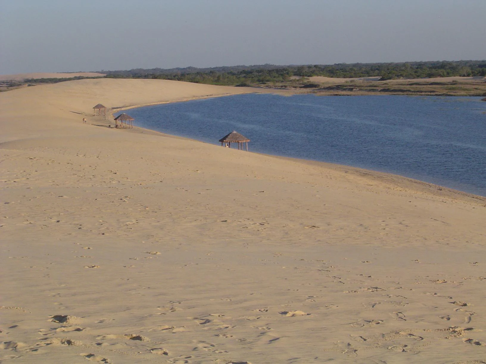 A wide angle view of expansive light tan sand dunes meeting a blue lagoon with several thatched-roof shelters along the shoreline.
