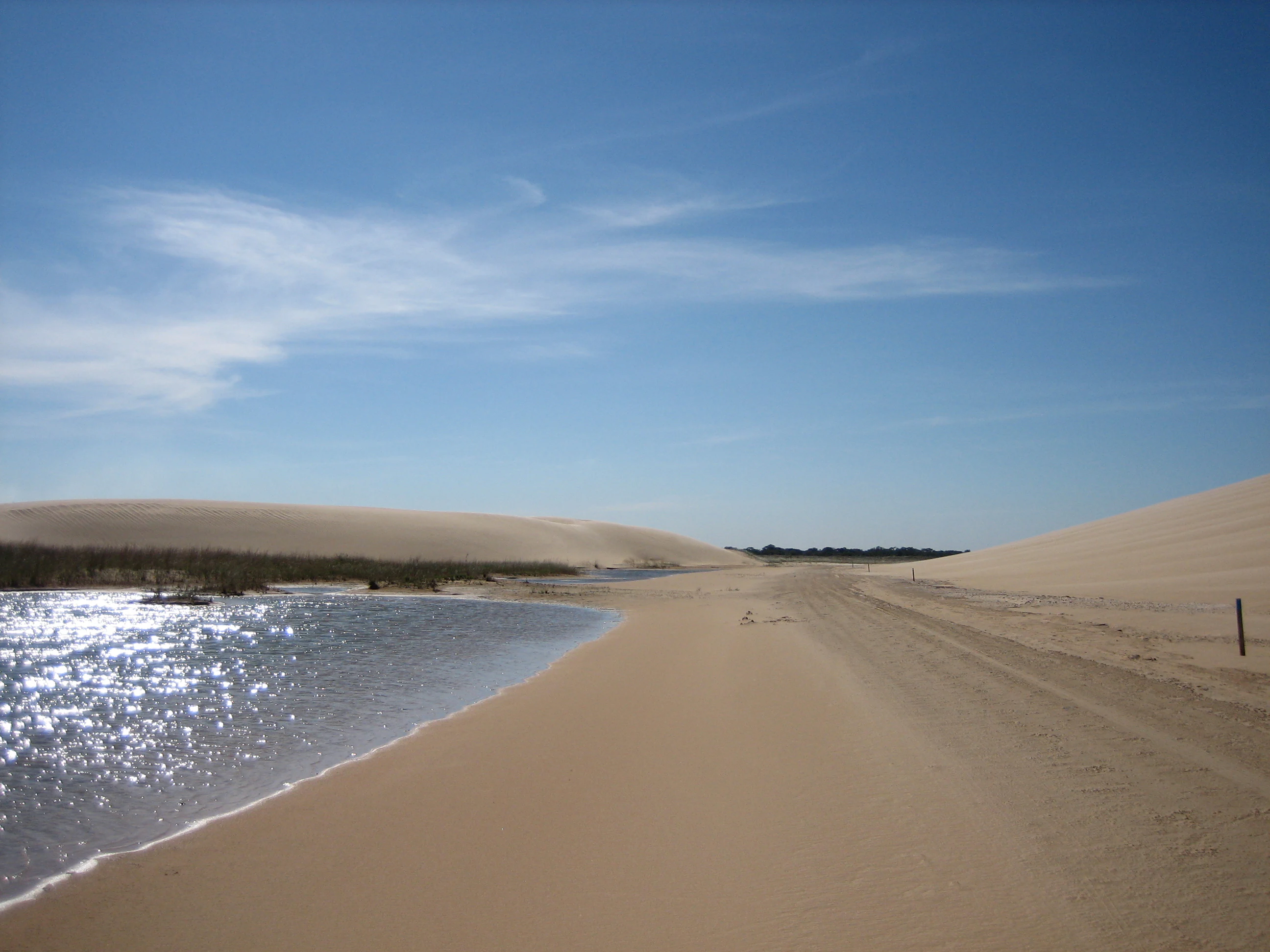 A wide view of light-colored sand dunes meeting a shallow lagoon under a bright blue sky with thin clouds.