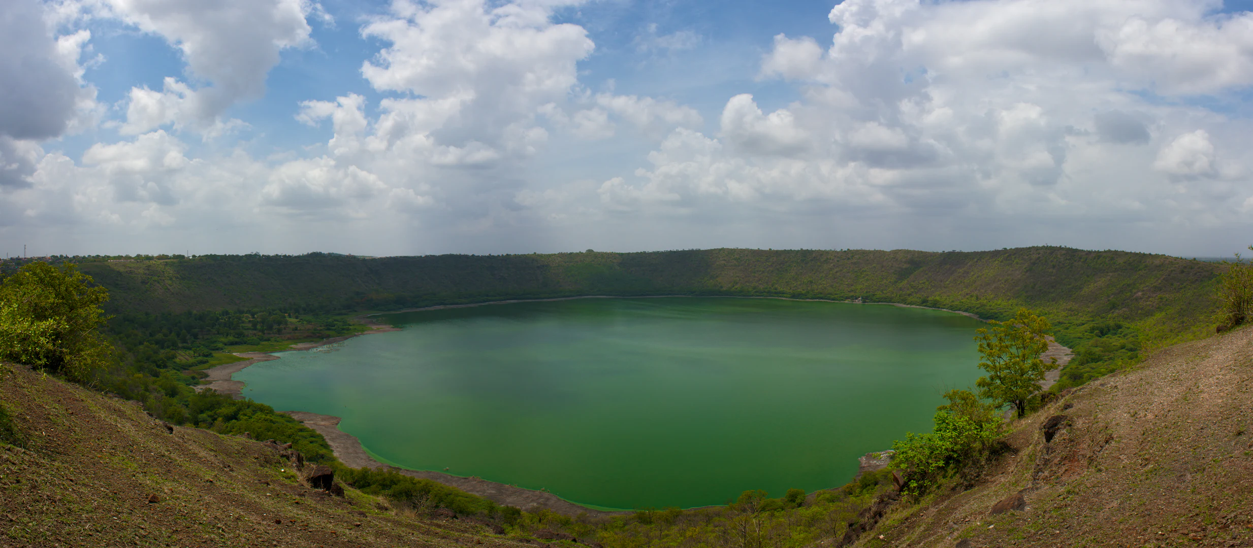 A wide, panoramic view looking down into the Lonar meteor impact crater, which contains a large, vibrant green circular lake surrounded by steep, vegetated crater walls under a cloudy sky.