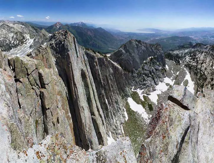 A wide view of a steep, craggy, light-colored granite mountain peak and cliff face with patches of snow remaining in the rocky basins below.