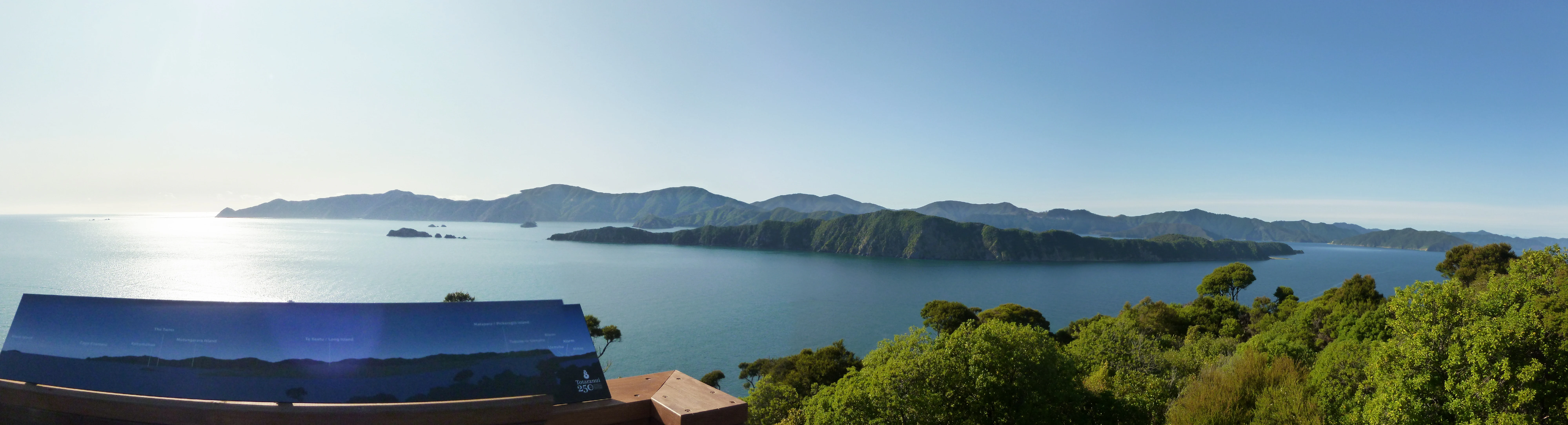 A wide panoramic photograph showing a calm body of water with a long, hilly, tree-covered island in the distance under a clear blue sky, viewed from a high vantage point with a dark interpretive sign in the foreground.