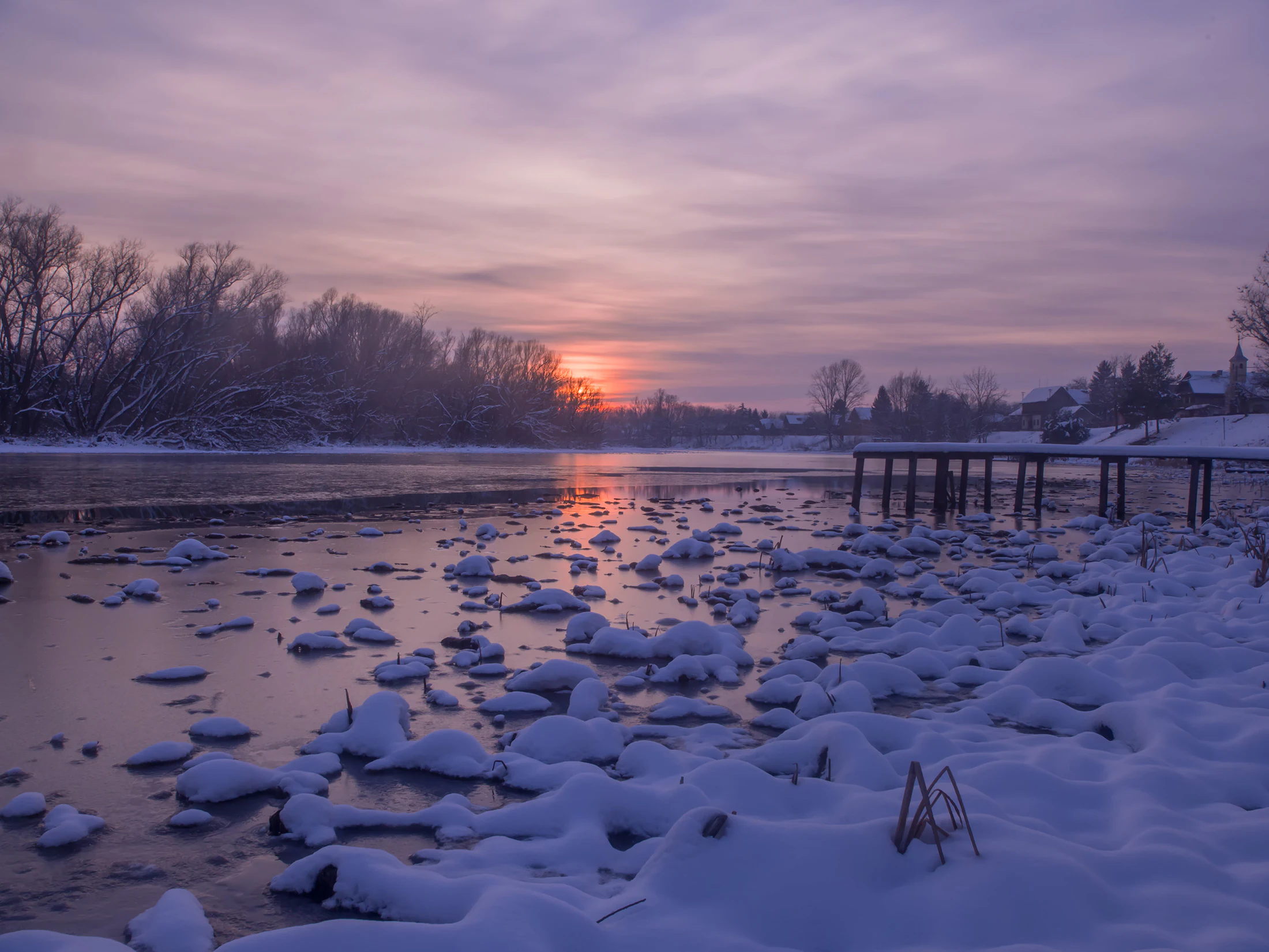 Snow-covered river with ice floes, wooden pier, and winter sunset sky