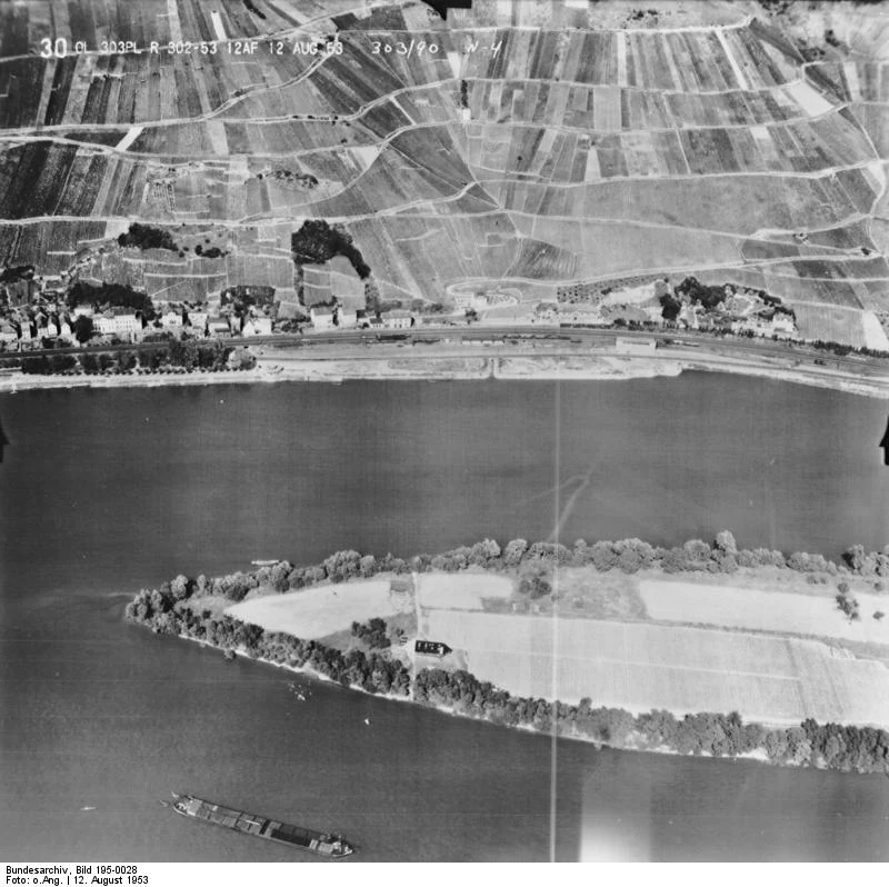 A black and white aerial photograph showing a section of the Rhine river with the Lorcher Werth river island in the foreground, and the town of Lorch with agricultural vineyard slopes in the background.
