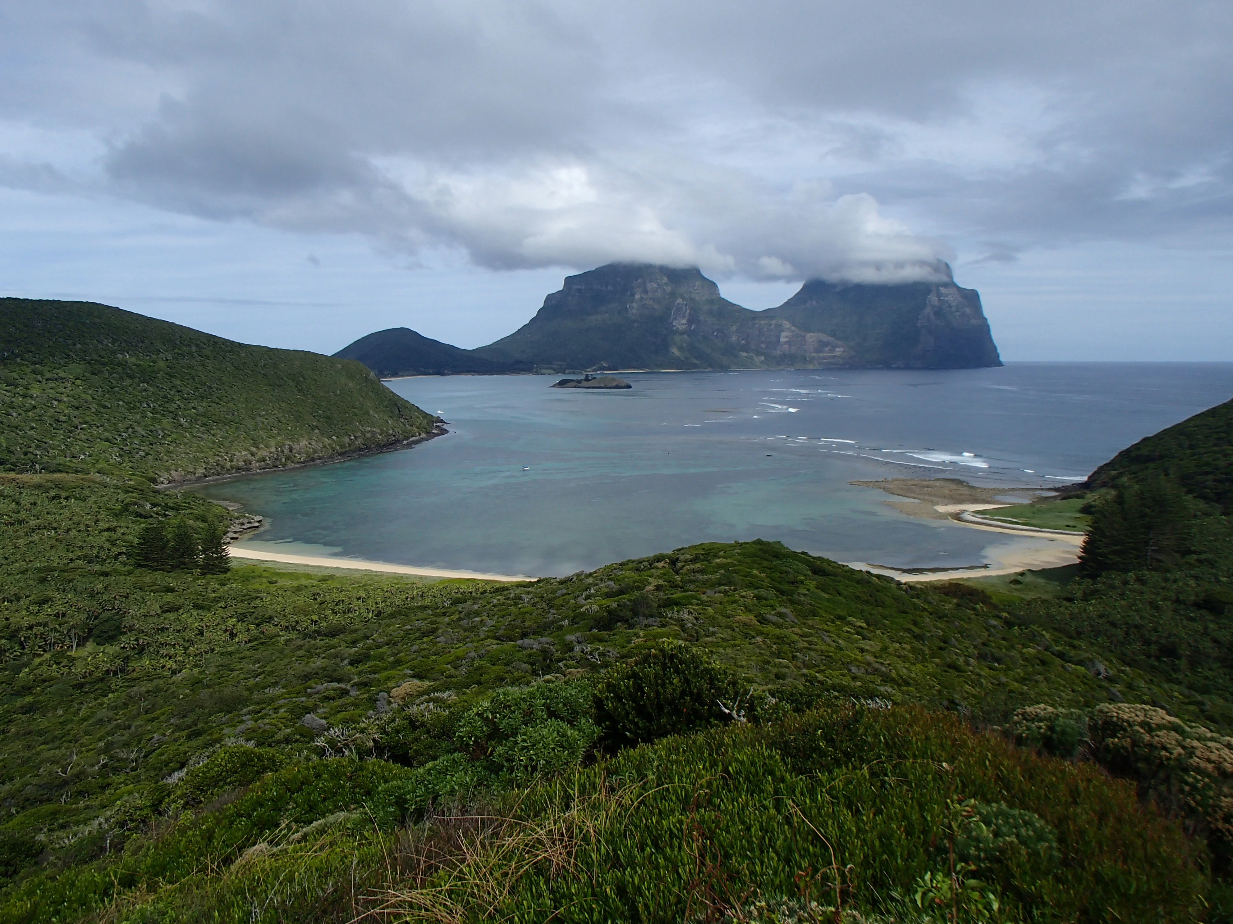 Wide landscape view overlooking a bay with turquoise waters and a sandy beach, with prominent mountains topped by clouds in the background.