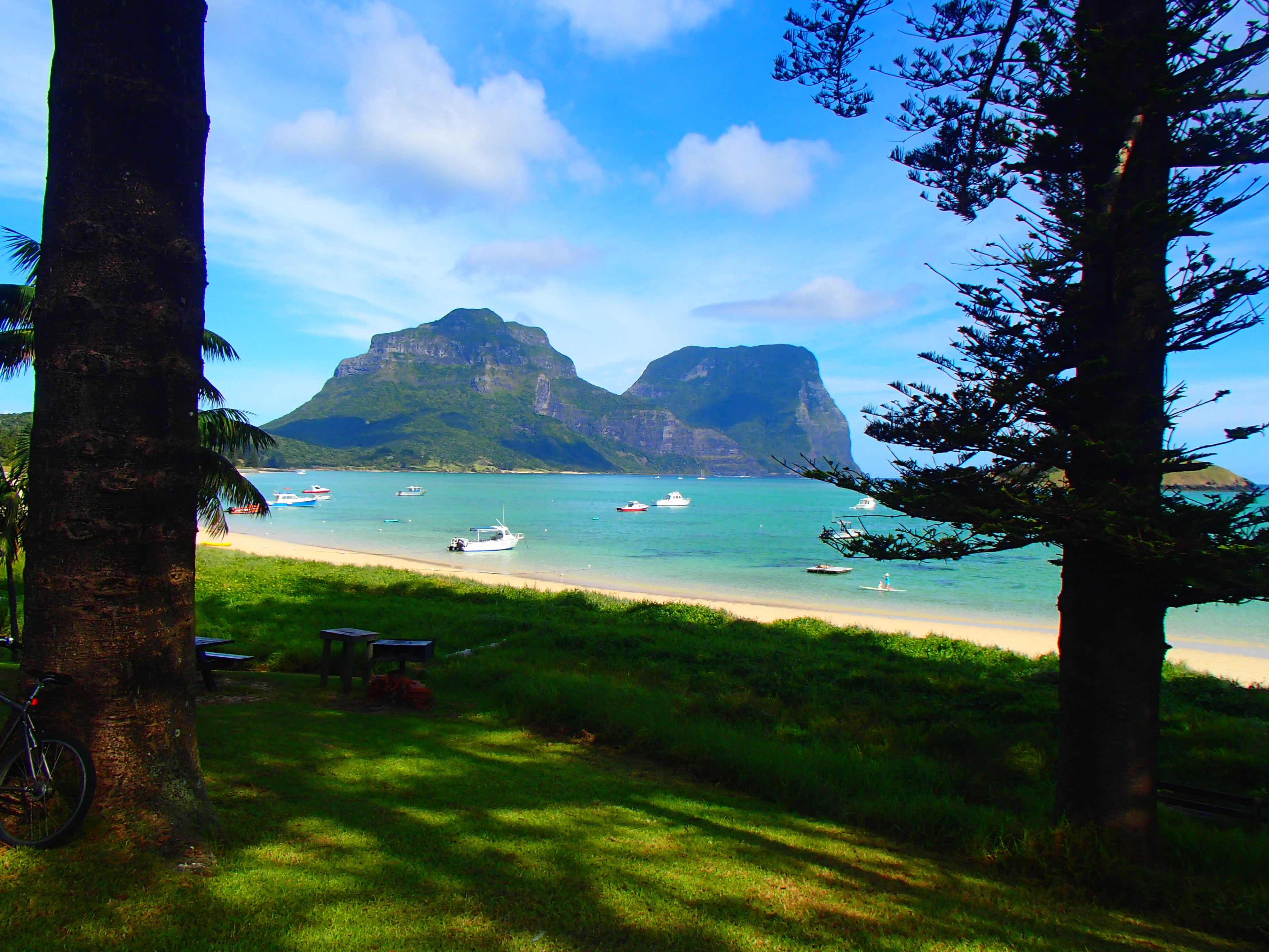 A view looking across a sandy beach and a turquoise lagoon toward two large, dark, rugged mountains framed by large pine trees.
