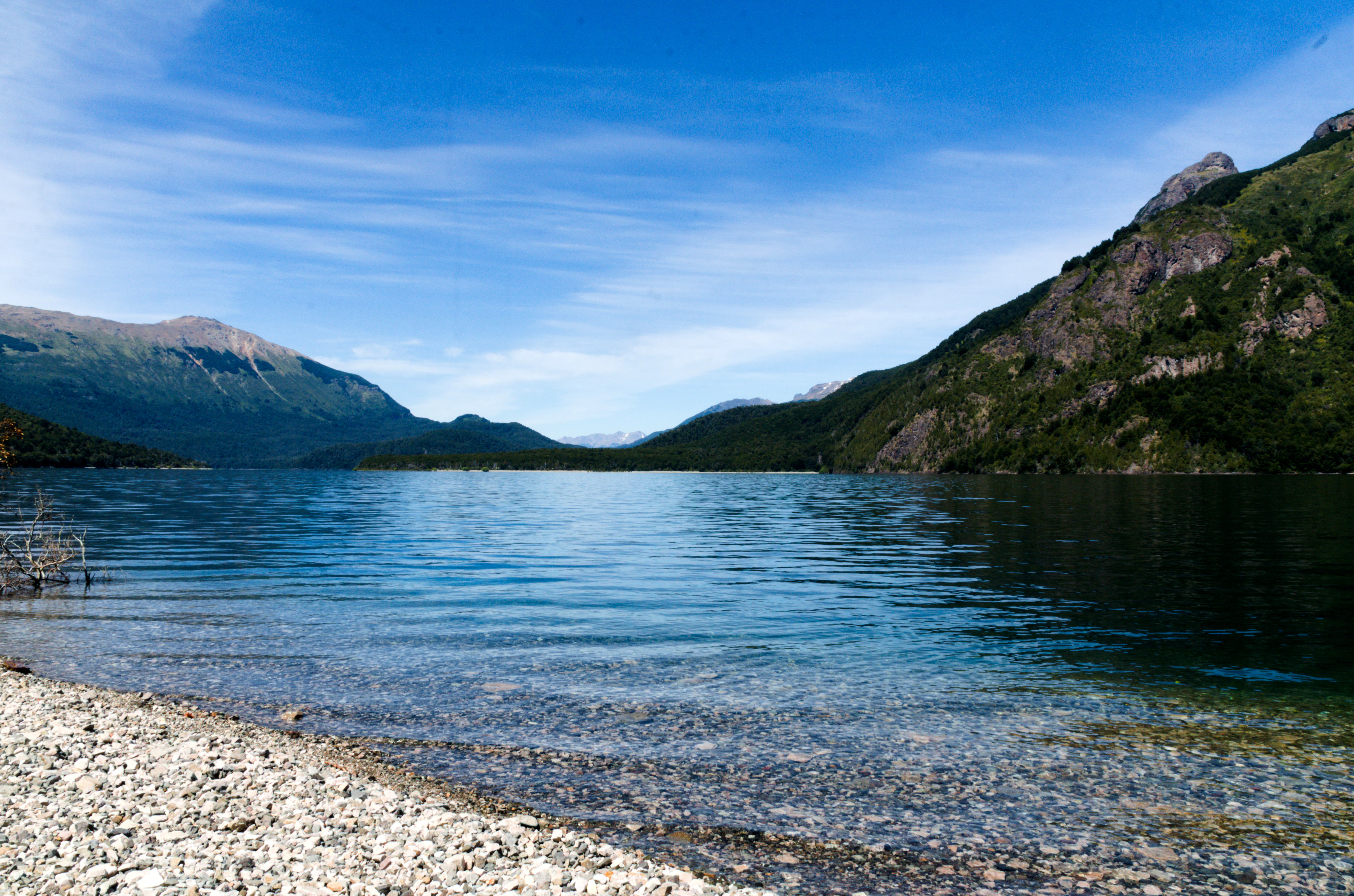Lake with clear blue water, rocky shore, and mountain backdrop under a clear blue sky.