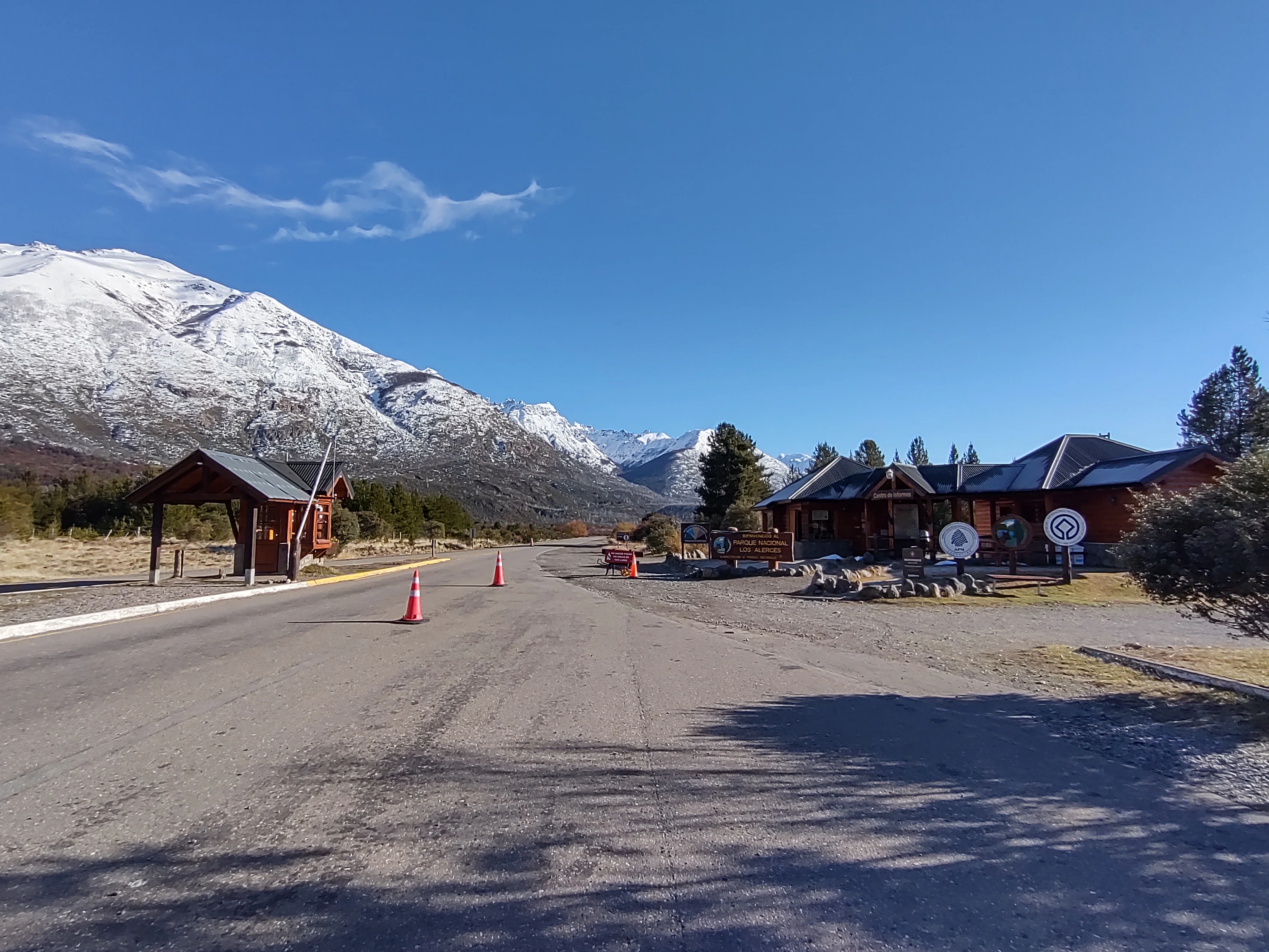 Road leading to park entrance with wooden structures, traffic cones, snow-covered mountains, and clear blue sky