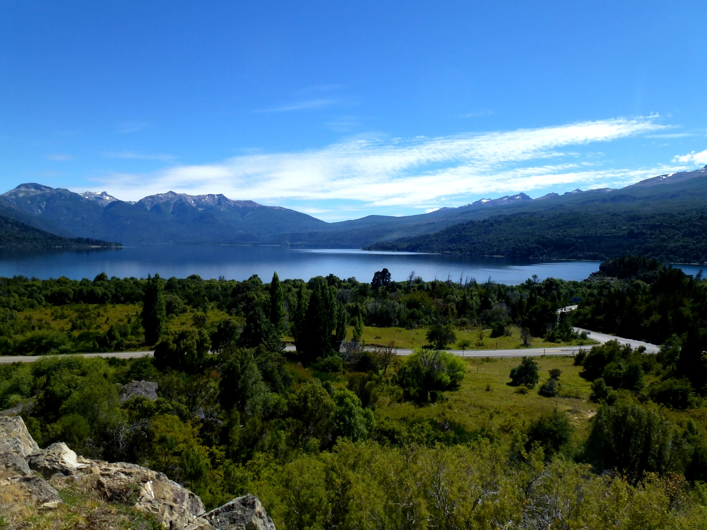 Lake surrounded by dense green forests and mountains under a clear blue sky with a road visible in the midground