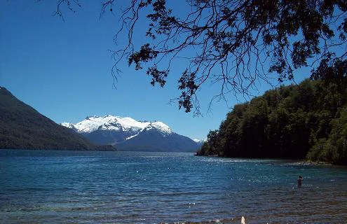 Lake Menendez with snow-covered mountains, forested shoreline, and a person wading in the water
