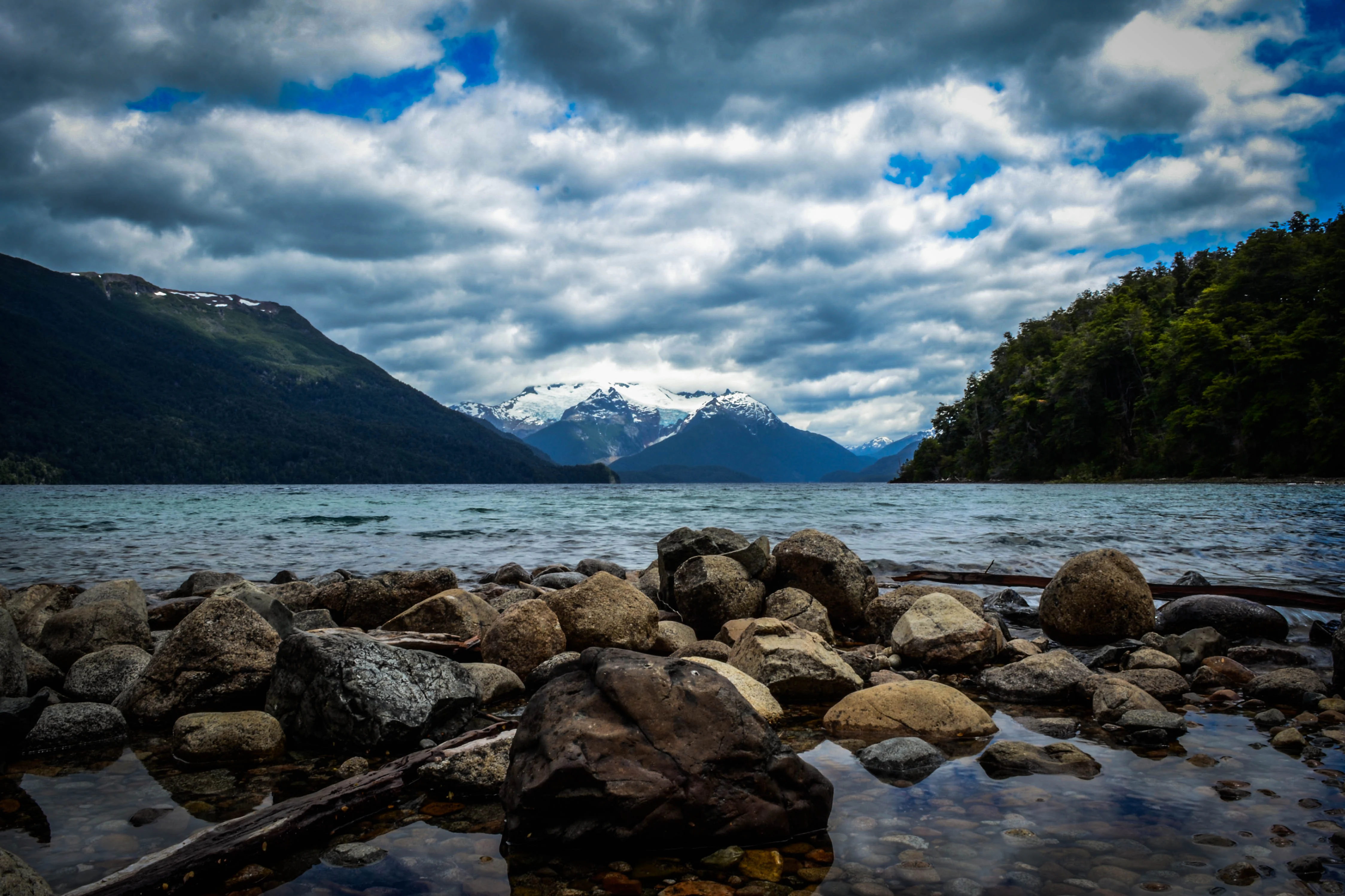 Lake with rocky shoreline, calm water, and snow-capped mountains under a partly cloudy sky