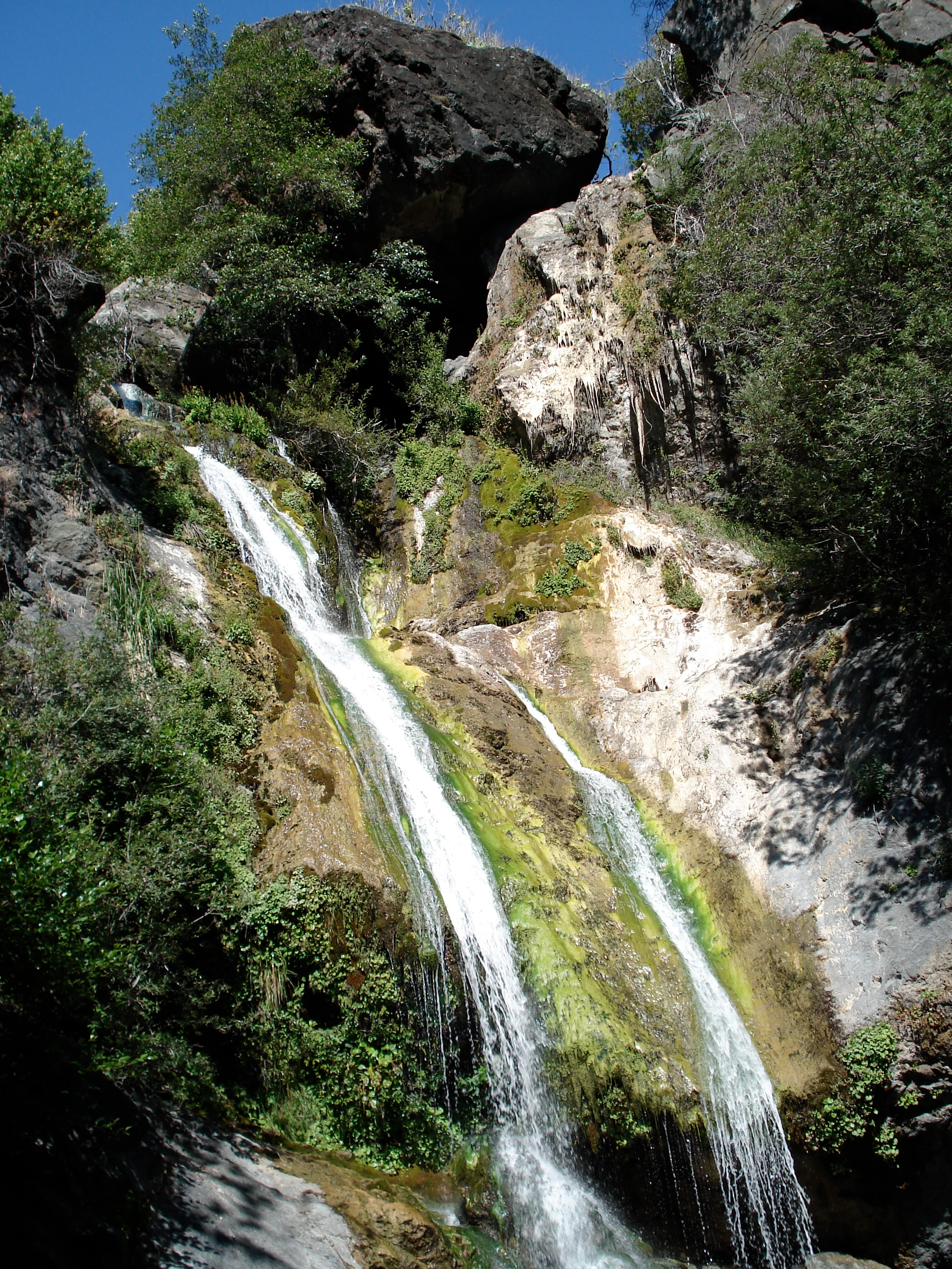 A multi-tiered waterfall flowing down a steep, mossy rock cliff face surrounded by dense green vegetation.