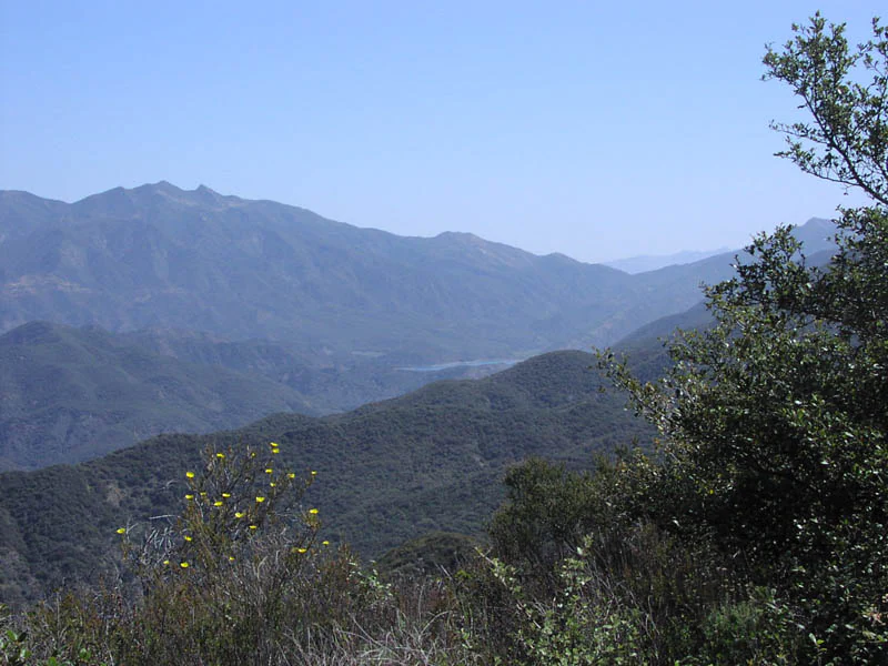 A wide view of layers of hazy mountain ridges under a clear blue sky, with a reservoir visible in the midground and chaparral vegetation in the foreground.