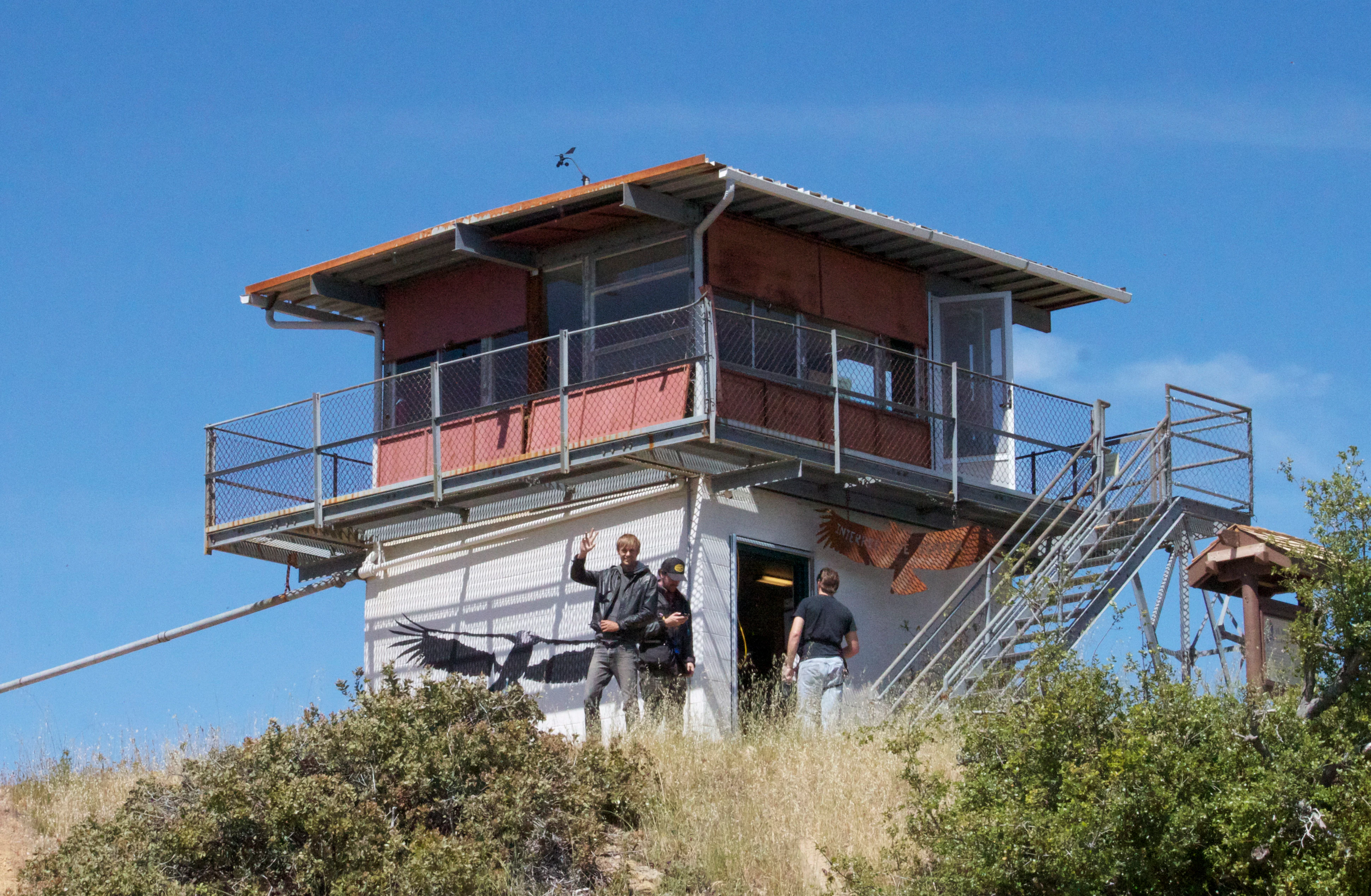 A square, elevated fire lookout building with a wrap-around balcony and external metal staircase standing on a grassy hill under a clear blue sky, with three people standing near the base.