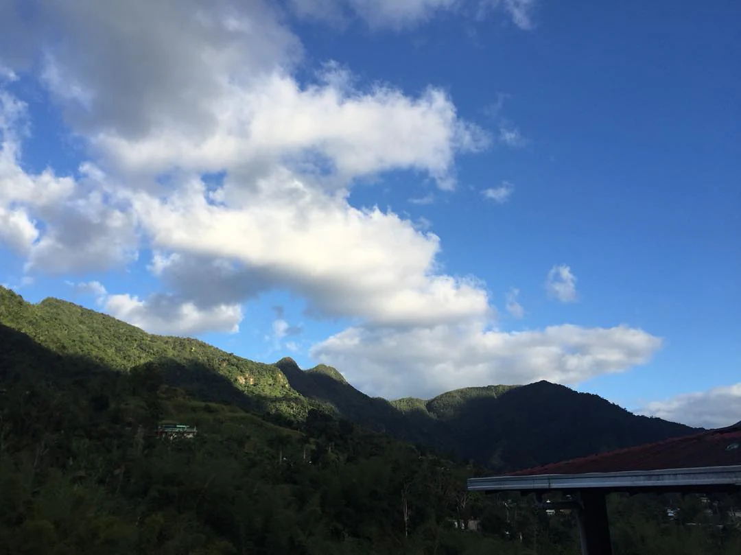 A wide mountain landscape featuring lush green peaks under a blue sky with scattered clouds, with a dark structure visible in the bottom right corner.