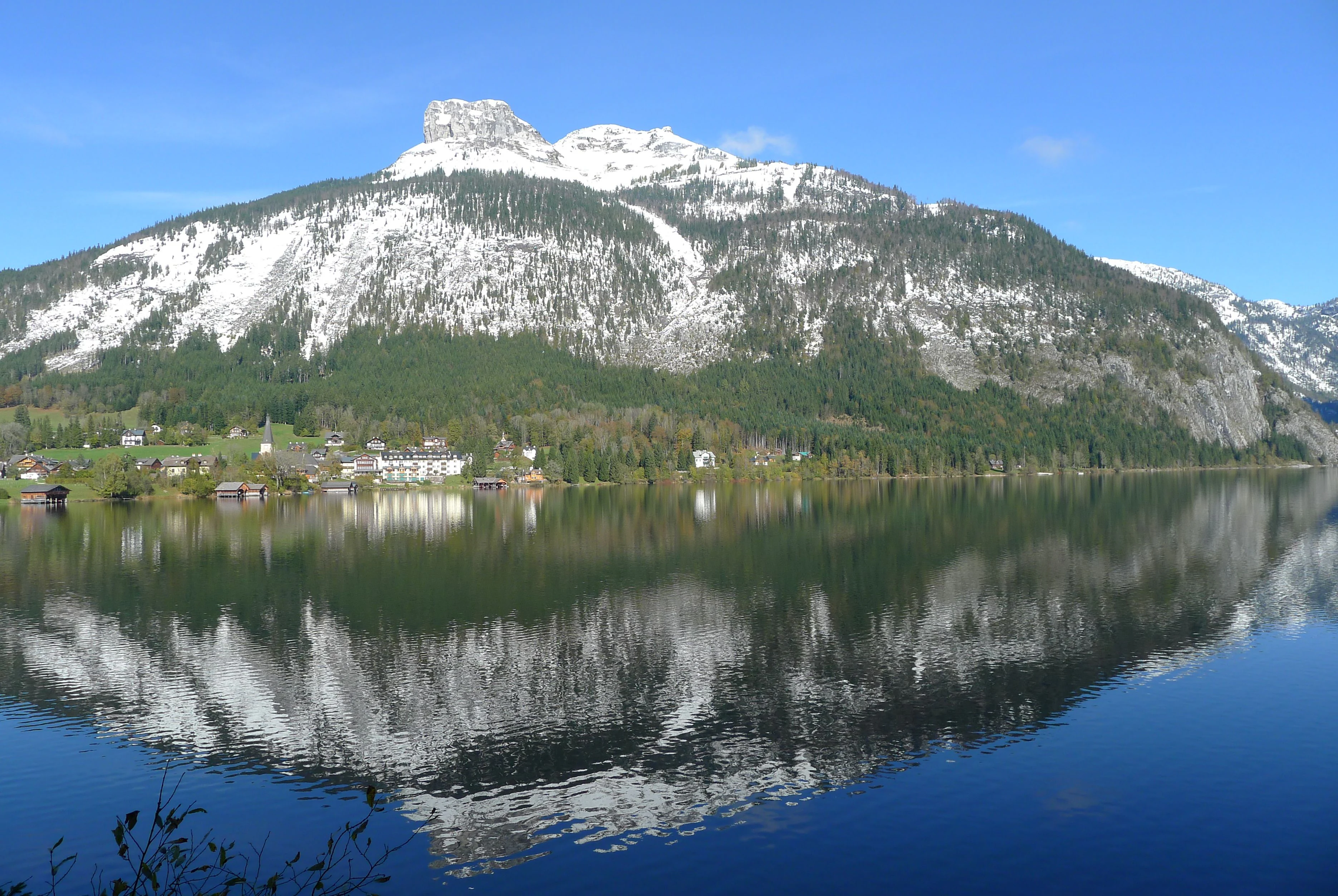 Una toma amplia a la altura de los ojos de una gran montaña nevada llamada Loser, que se refleja claramente en el agua oscura y tranquila del lago Altaussee bajo un cielo azul brillante.