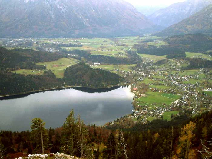 Una vista amplia de estilo aéreo que mira hacia abajo desde una cresta montañosa alta hacia un lago alpino oscuro, un pequeño pueblo con campos verdes y laderas boscosas circundantes.