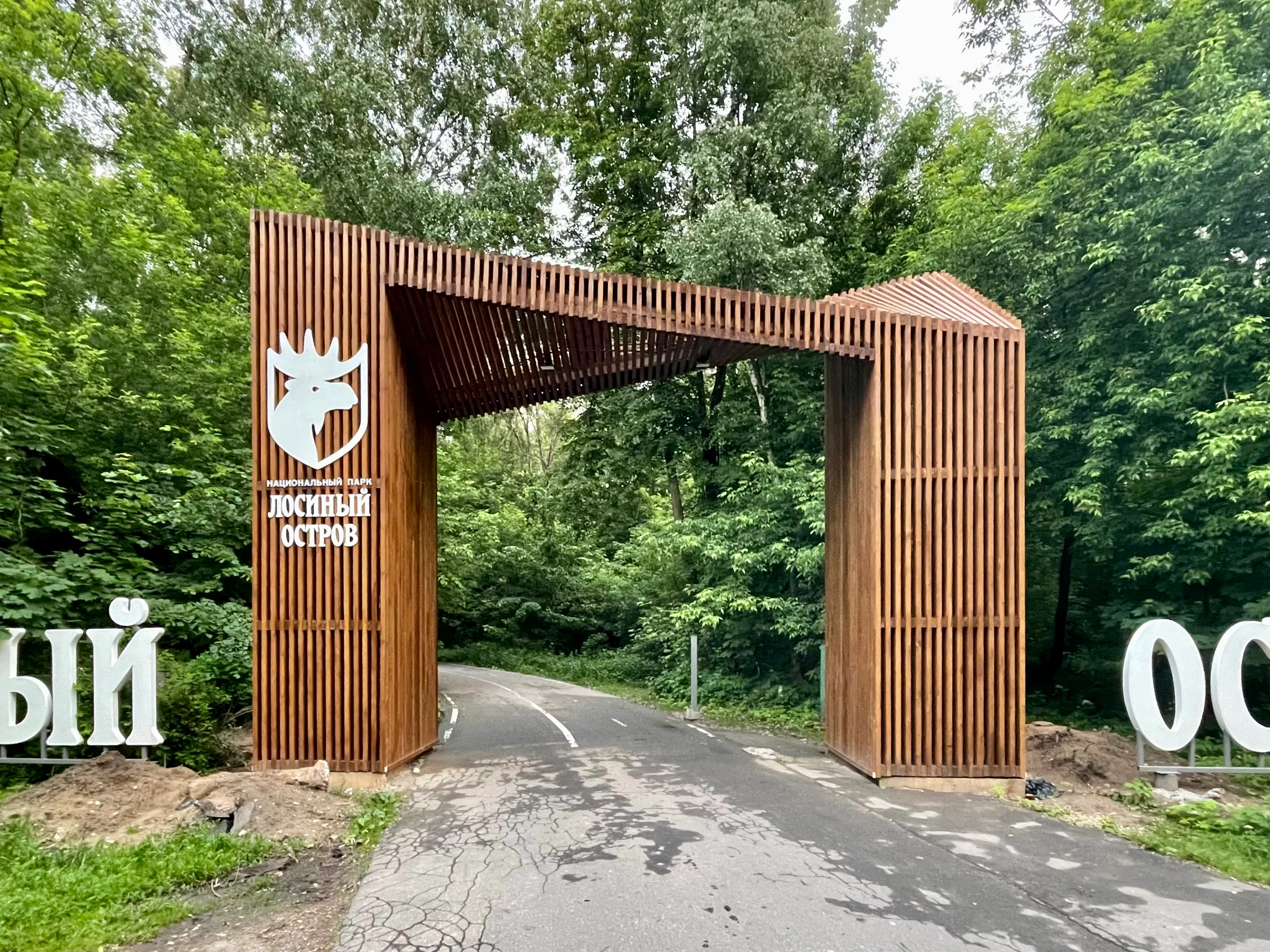 Una gran puerta de entrada de madera con listones verticales cruza una carretera asfaltada que se adentra en un bosque. El pilar izquierdo muestra el logotipo de una cabeza de alce blanca y el texto 'ЛОСИНЫЙ ОСТРОВ'. Grandes letras blancas son parcialmente visibles en el suelo a ambos lados de la puerta.