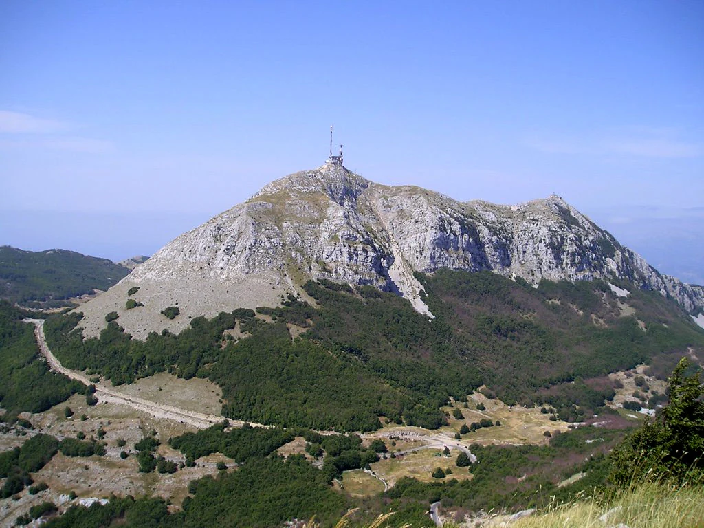A wide view of a rocky mountain peak rising above a forested landscape under a clear blue sky, featuring a communications tower at the summit.