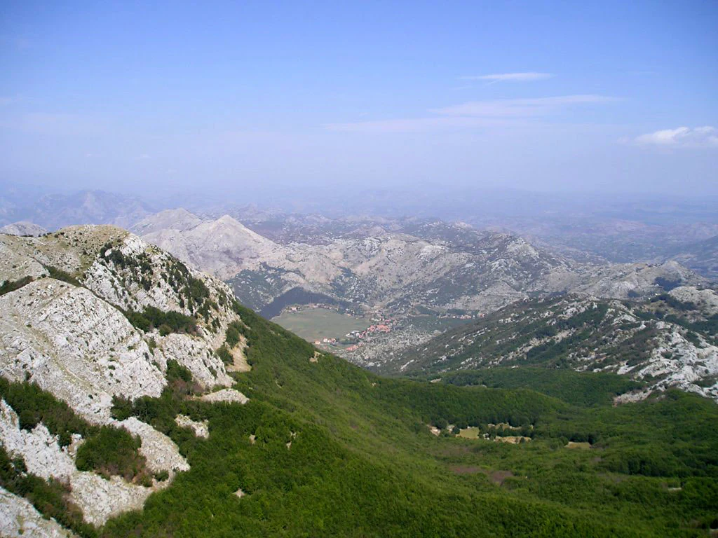 A wide, elevated view of rugged, rocky limestone mountains descending into lush green valley slopes in Montenegro.