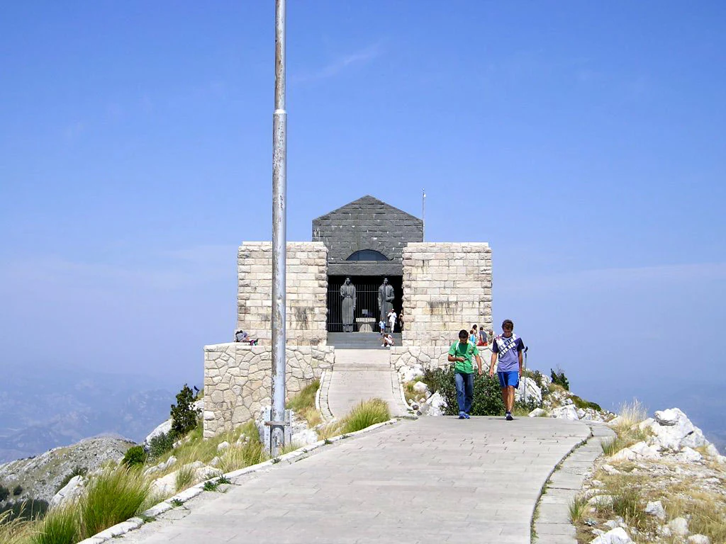 Stone mausoleum structure with people walking on a paved path toward it under clear blue sky