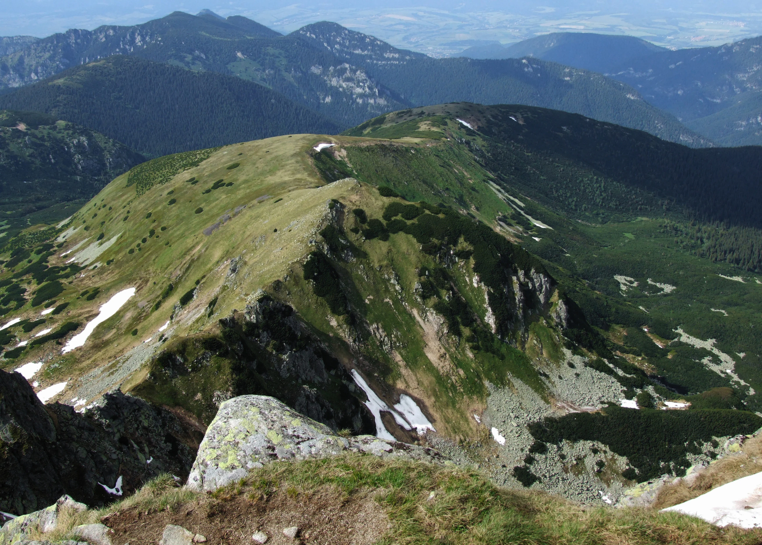 Grassy mountain ridge with patches of snow, rocky outcrops, and distant mountain ranges under clear sky