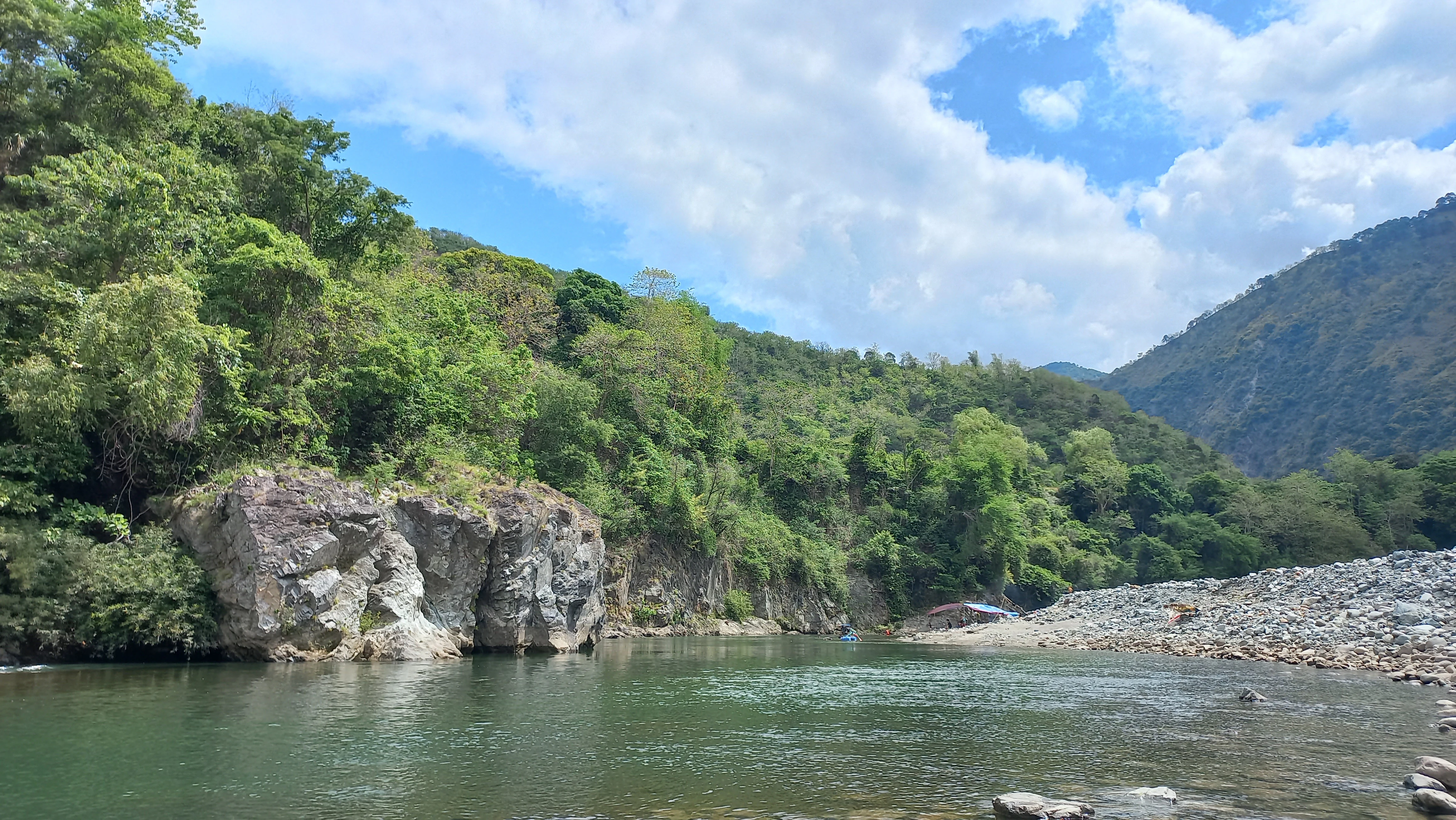 A green, tree-covered mountainside rises above a calm river with a prominent rocky schist outcrop on the left bank and a stony riverbed on the right under a blue sky with white clouds.