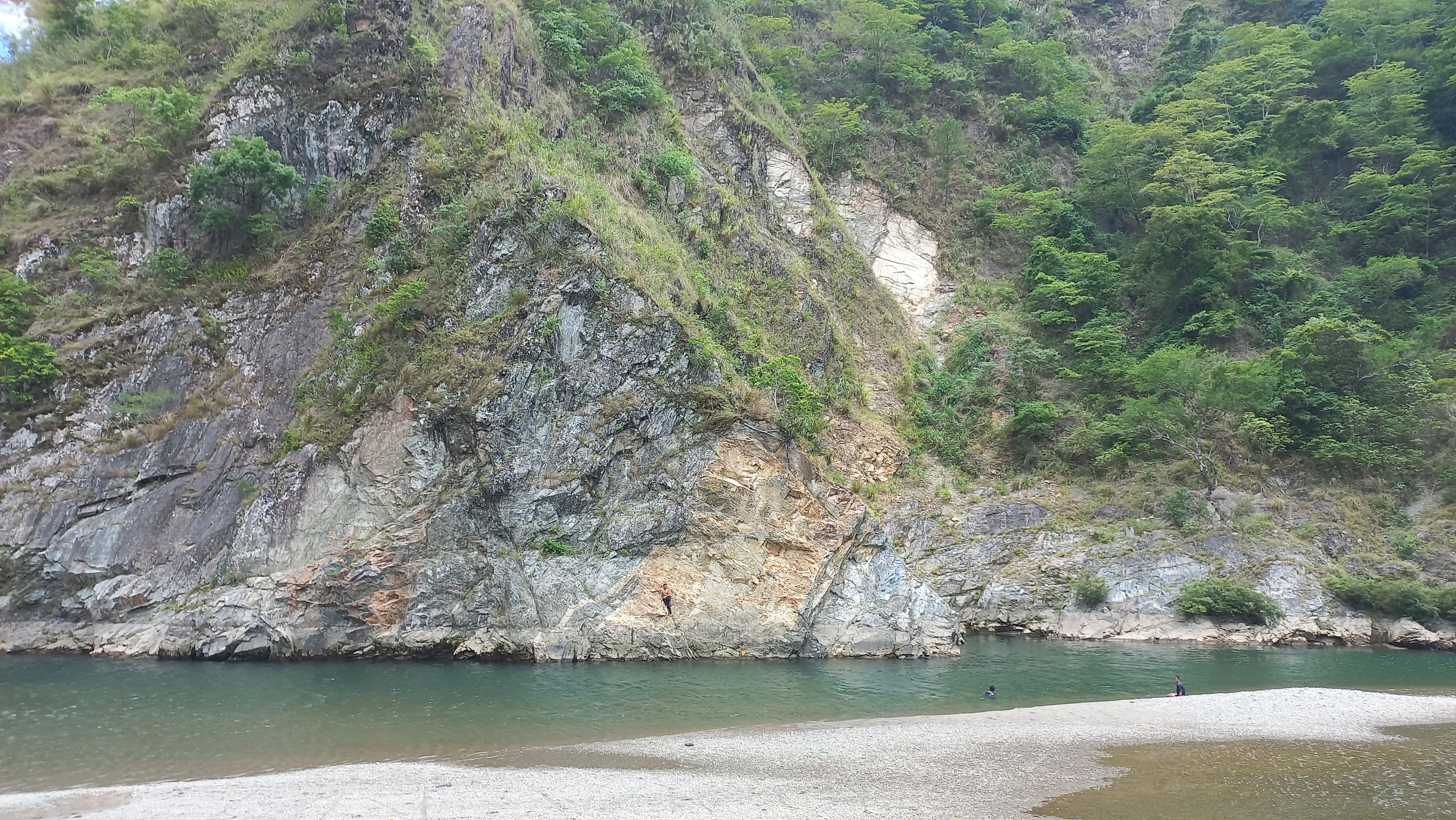 A steep, rocky riverbank with exposed schist and a prominent light-colored diorite dike, viewed across the Agno River.