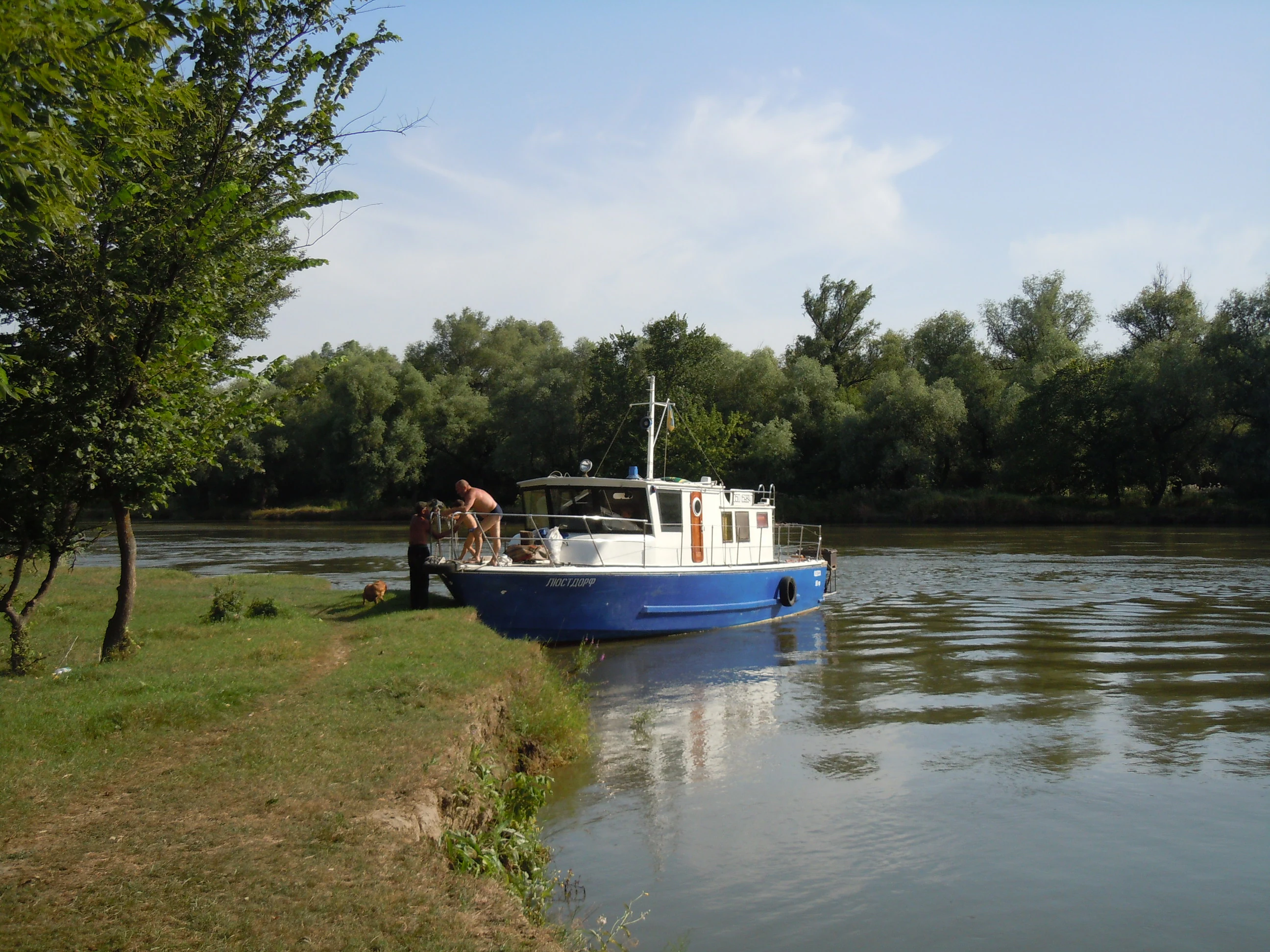Blue and white boat docked on riverbank with grassy shore, trees, and two people nearby