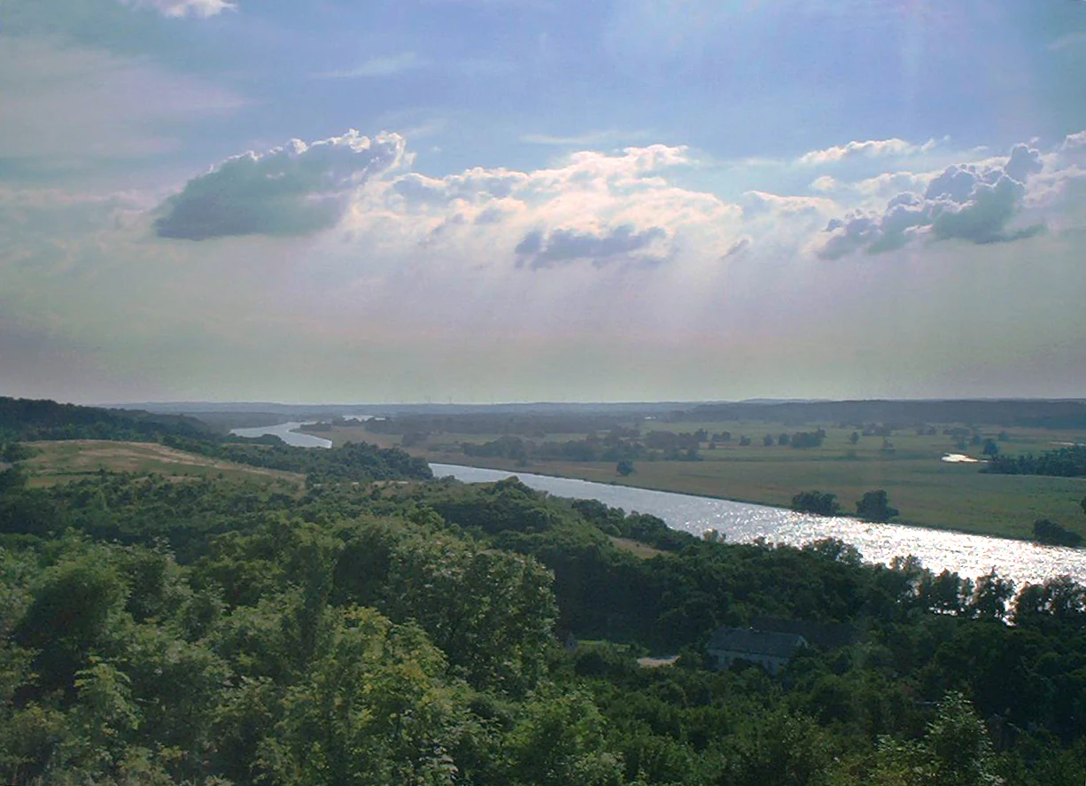 Wide landscape view of the Oder River flowing through lush green floodplain with rolling hills under a partly cloudy sky.