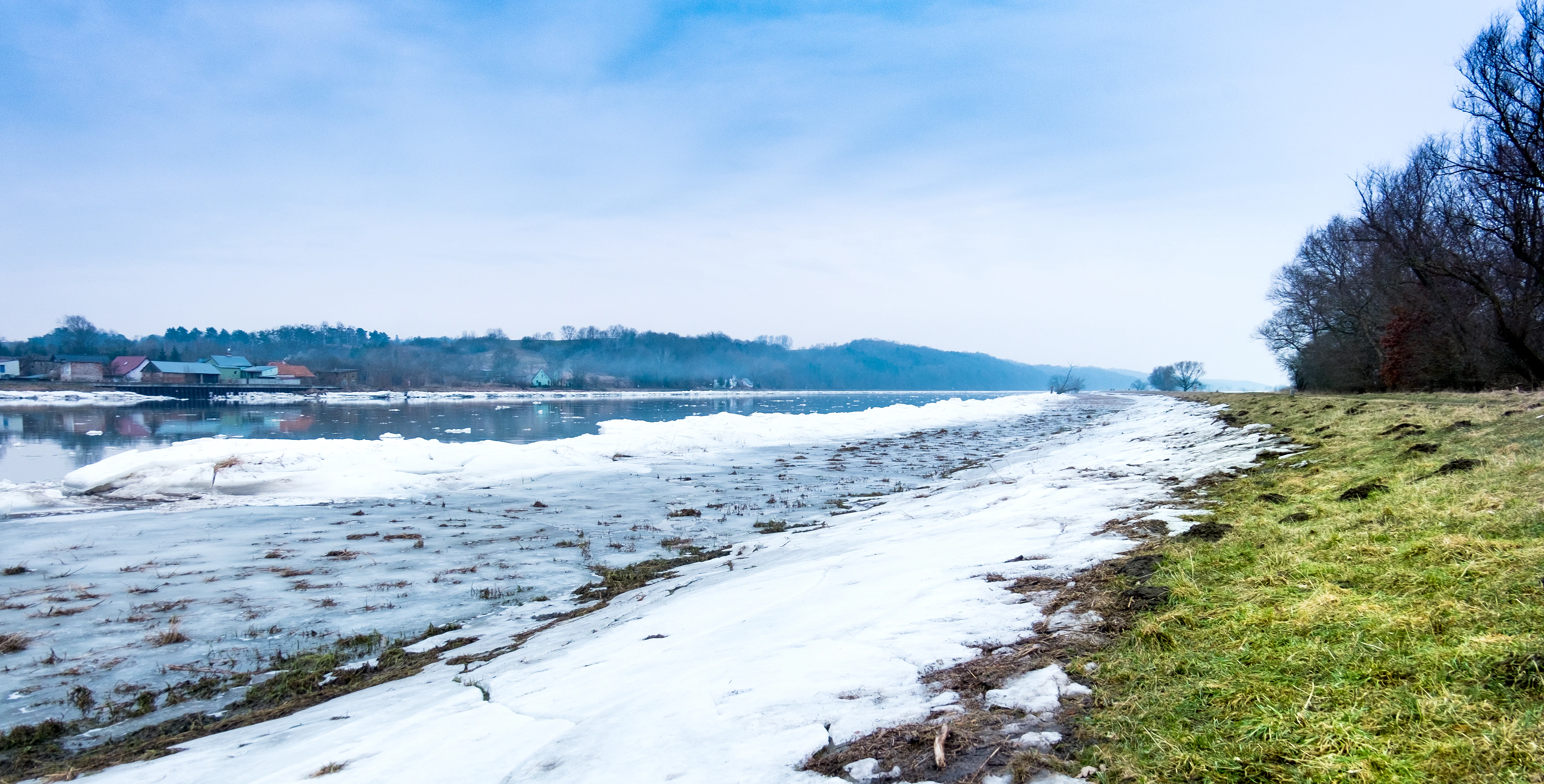 River Oder with snow-covered bank, grassy shoreline, and distant hills under a clear sky