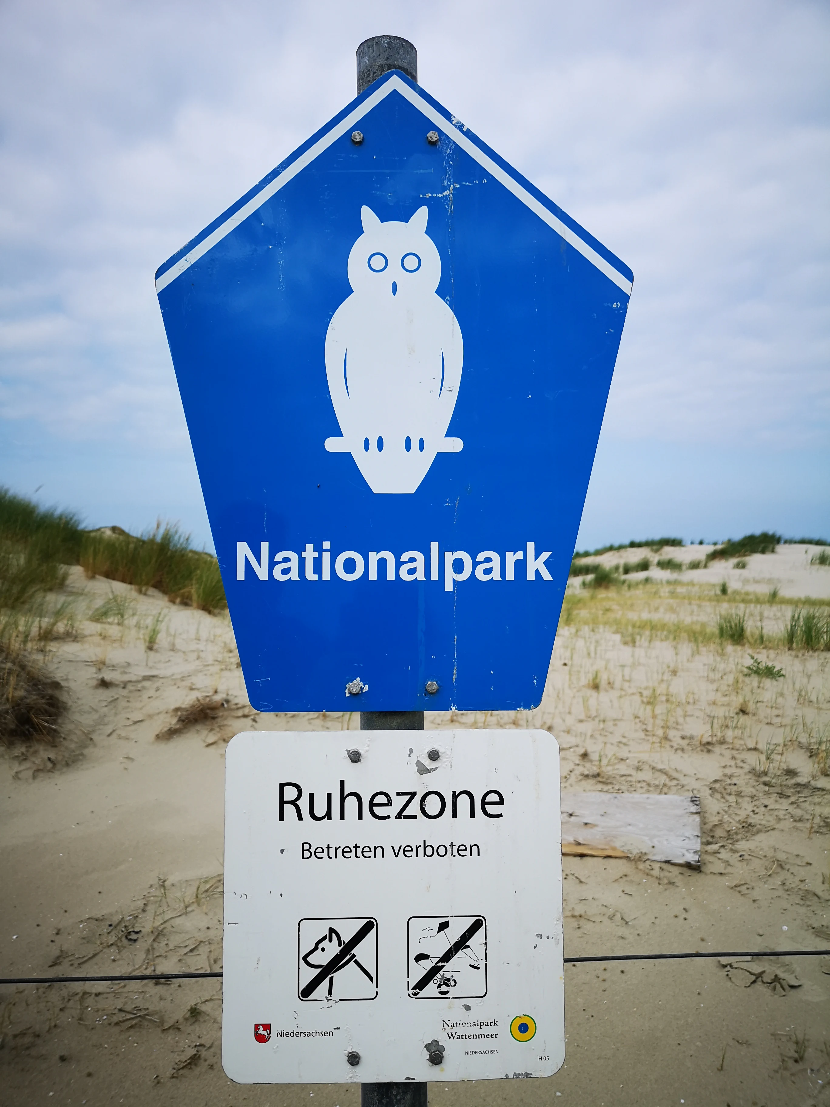 Blue sign with white owl symbol and 'Nationalpark' text, mounted above a white sign reading 'Ruhezone' with prohibited activity symbols, set against sandy dunes and grassy background