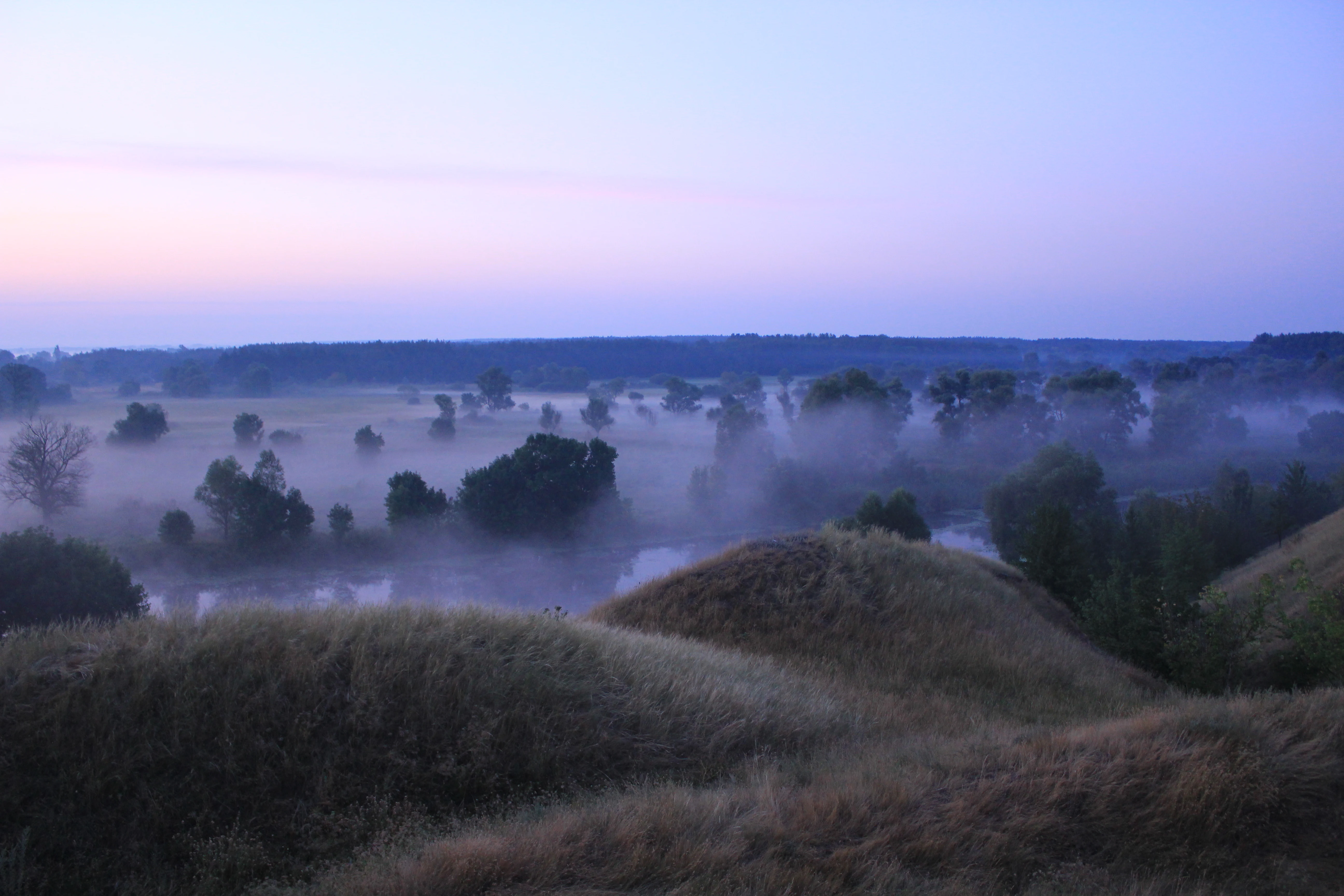 Misty landscape with rolling hills, trees, and a river in the distance under a dawn sky