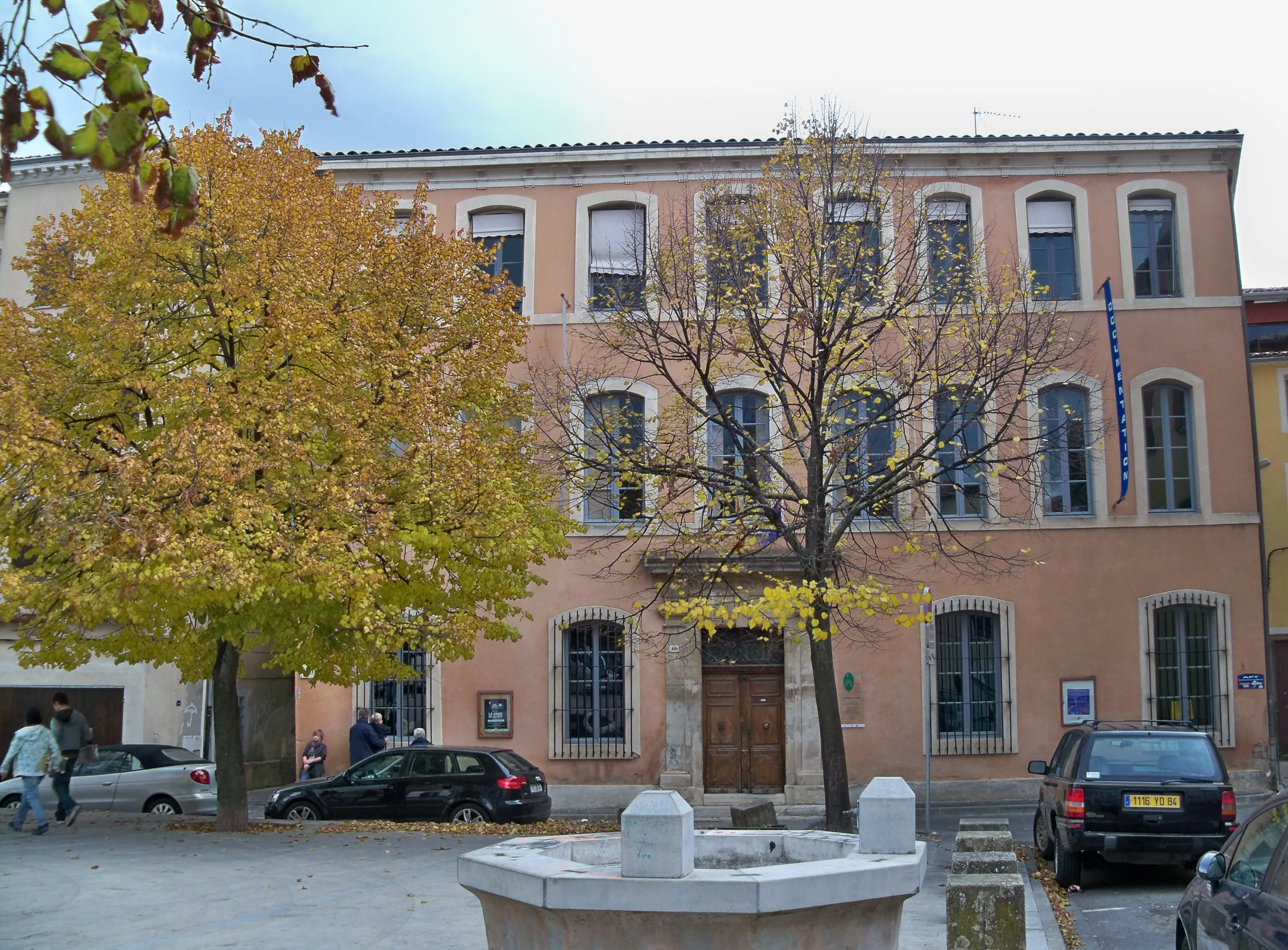 Un edificio de varios pisos de color melocotón con grandes ventanales y una puerta de madera, situado en una plaza con árboles de follaje otoñal, coches aparcados y una pequeña fuente de piedra en primer plano.