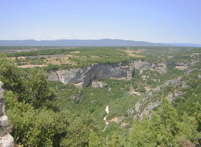 Una vista panorámica de gran angular que mira hacia un valle boscoso con acantilados de piedra caliza desde un punto elevado, con una cadena montañosa brumosa visible en el horizonte.
