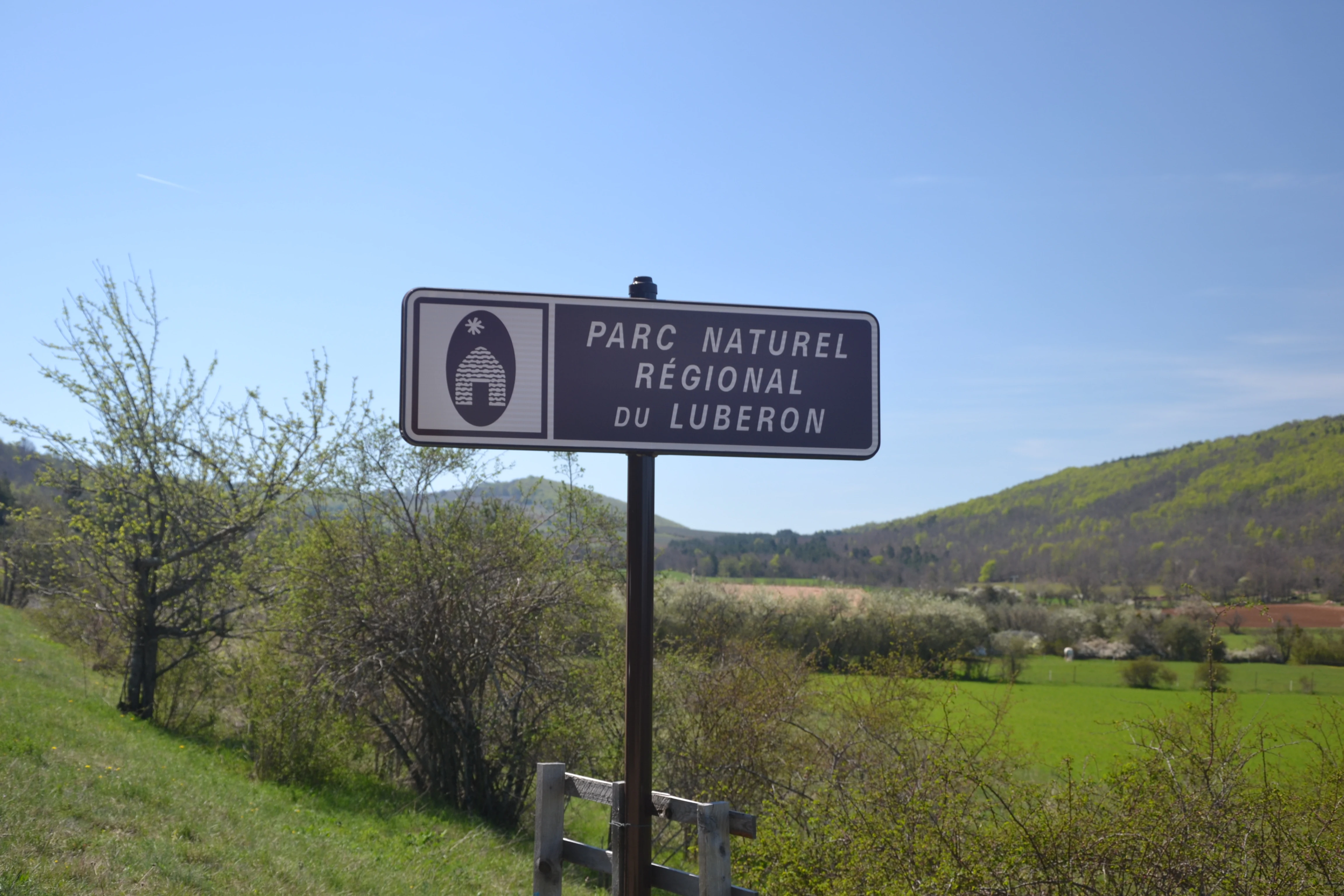 Una señal de carretera de metal marrón indica Parc naturel régional du Luberon, con un fondo de campos verdes y colinas onduladas bajo un cielo azul despejado.
