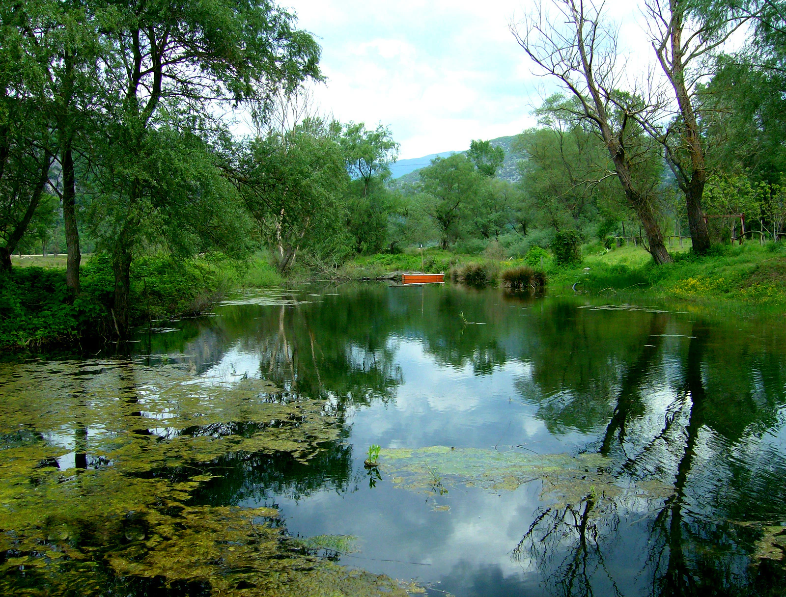 A calm section of the Nestos River reflecting trees and a cloudy sky, with a small orange boat docked near the far bank.
