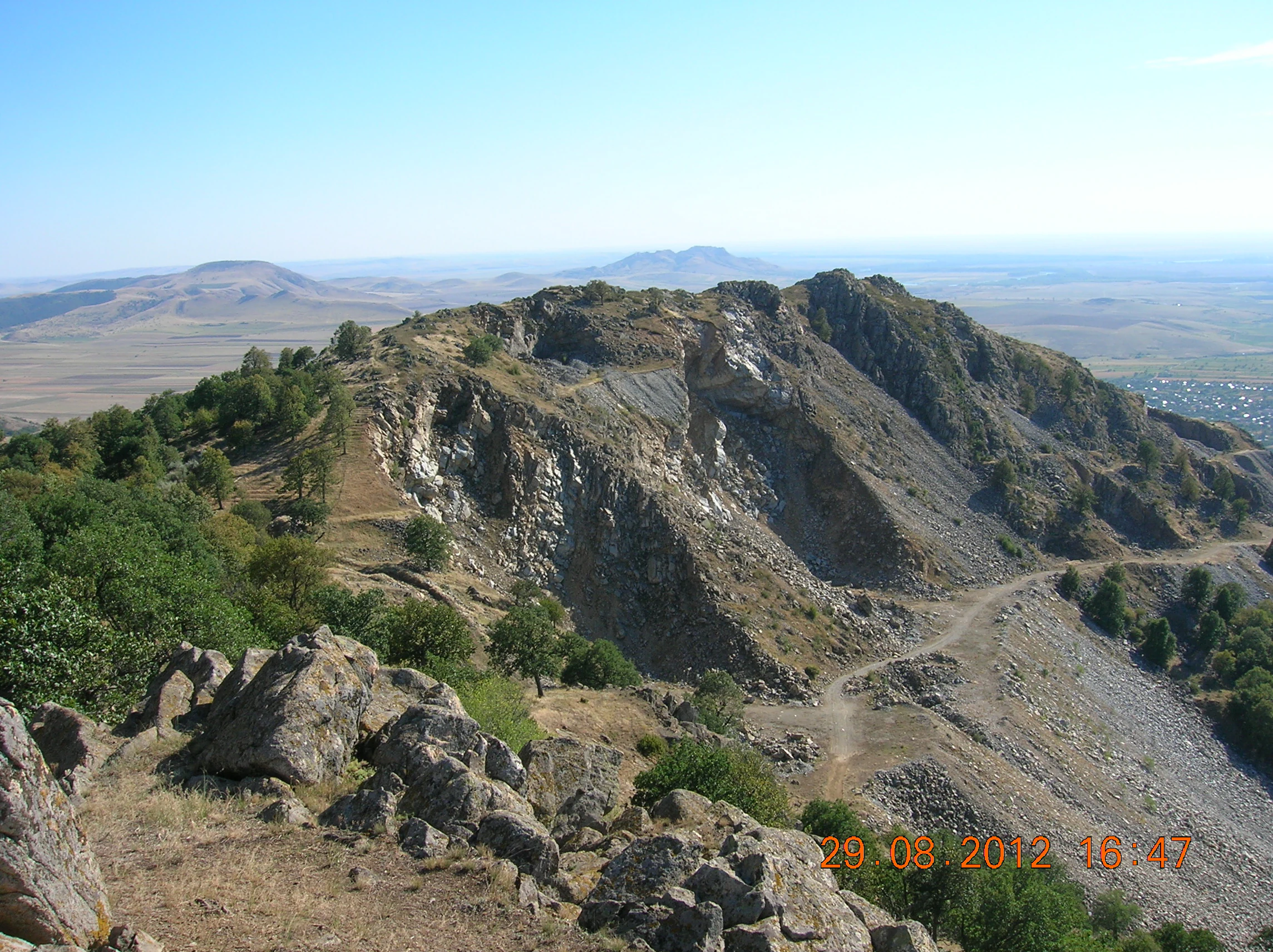 Rocky mountain peak with scattered trees and a winding path overlooking a vast plain under clear blue sky