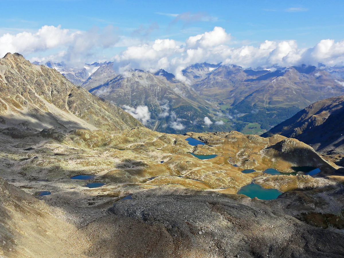 A wide aerial view of the Macun plateau in the Swiss Alps, showing numerous small, turquoise glacial lakes scattered across a rocky, high-alpine landscape with surrounding mountain peaks under a partly cloudy sky.