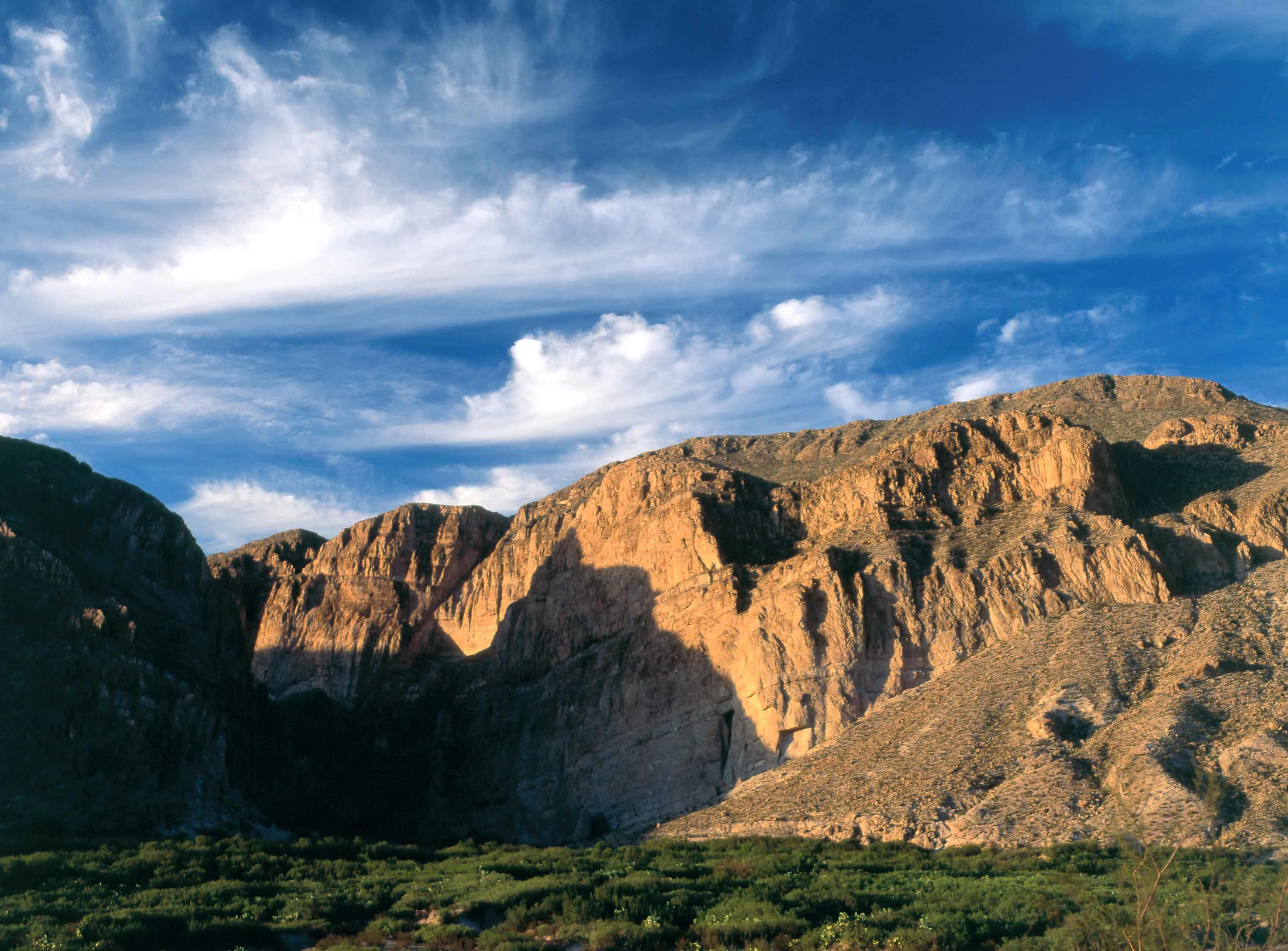 A wide landscape view of dramatic, sunlit rocky desert mountains under a blue sky with wispy clouds, with a foreground of green desert shrubbery.