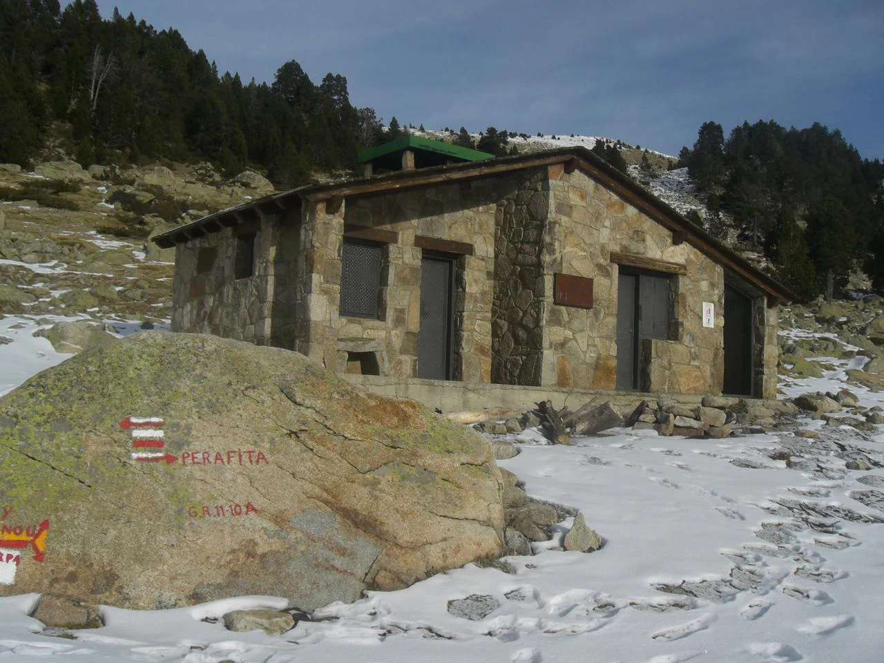 A rustic stone cabin, known as Perafita Refuge, sits on a snow-covered alpine slope with a large rock in the foreground marked by red and white trail signs.