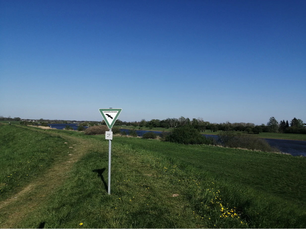 A triangular nature reserve sign on a metal post standing in a grassy meadow with the Weser river in the background under a clear blue sky.