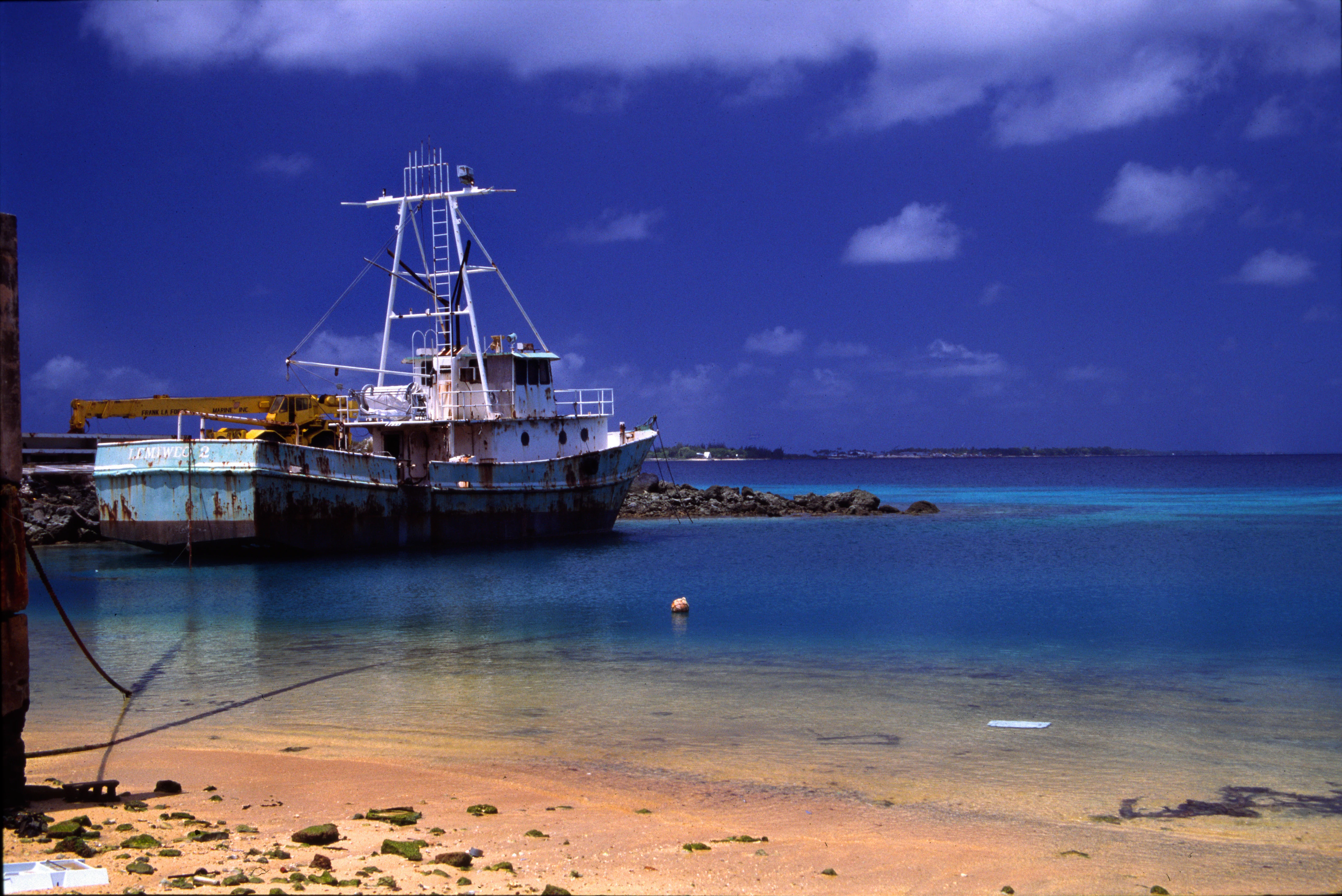 A rusted, white and blue fishing boat anchored in shallow, clear blue lagoon water near a sandy beach under a dark blue sky.