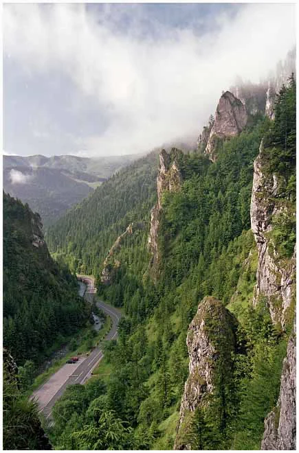 A mountain gorge with a winding road through dense forest, rocky cliffs, and clouds in the sky.