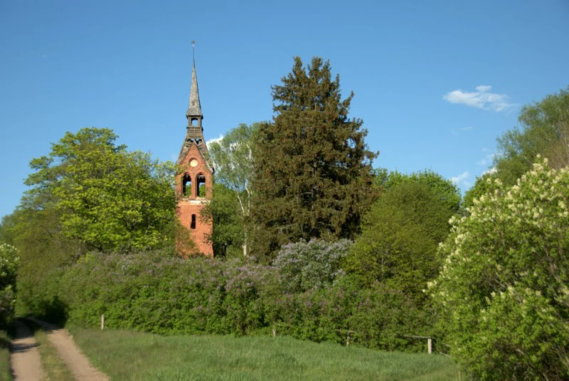 A red brick church tower stands partially obscured by trees and lush vegetation in a rural nature reserve, with a dirt path in the foreground.