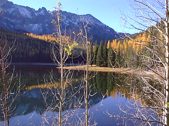 A calm lake reflects a mountain range and forest with autumn colors, viewed through bare tree branches in the foreground.