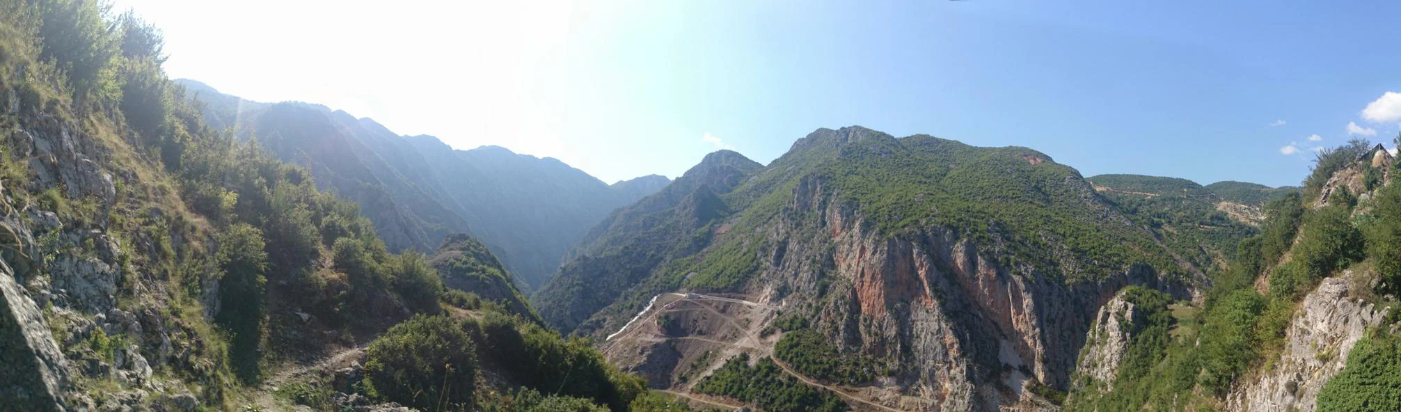 Panoramic view of a mountainous canyon with steep rocky slopes covered in sparse green forest vegetation under a clear sky.